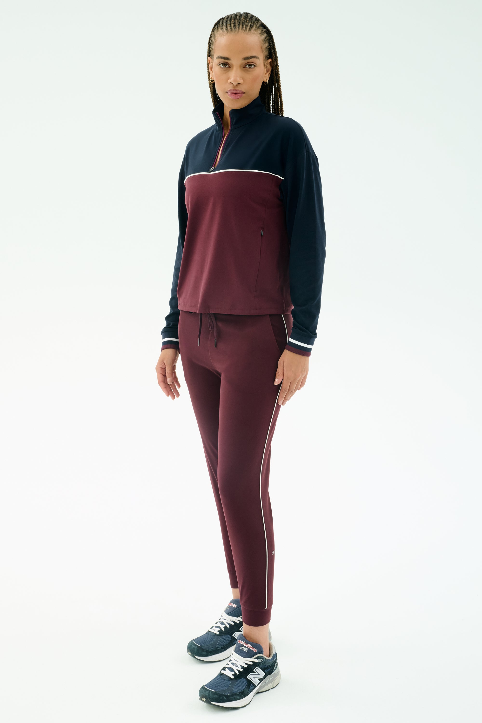 A woman faces forward against a white background, wearing the SPLITS59 Millie Airweight Half Zip in Dark Cherry/Indigo with navy sneakers and nylon-spandex leggings.