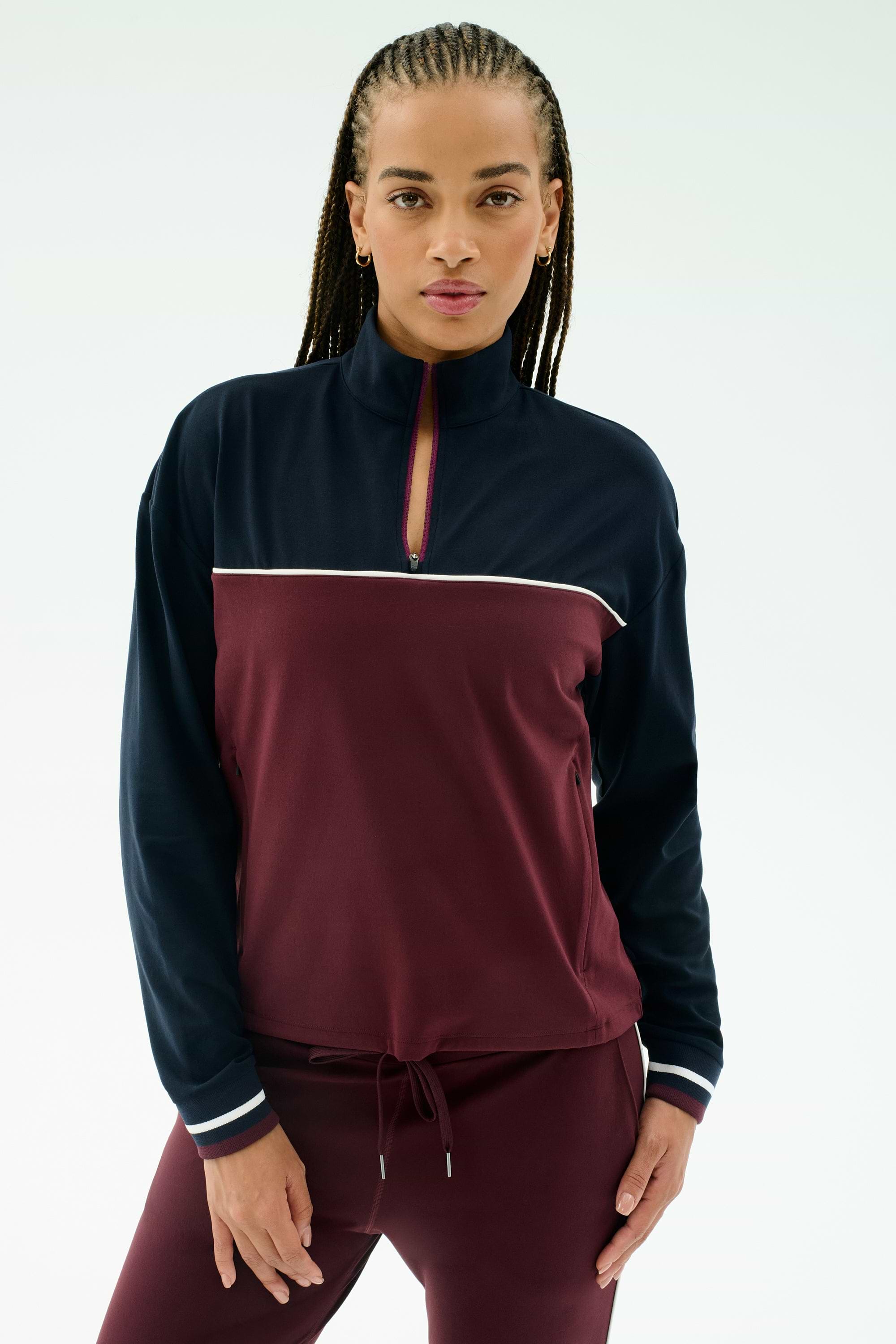 A woman wears the SPLITS59 Millie Airweight Half Zip in Dark Cherry/Indigo with matching pants, made from a nylon-spandex blend, standing against a plain light background.
