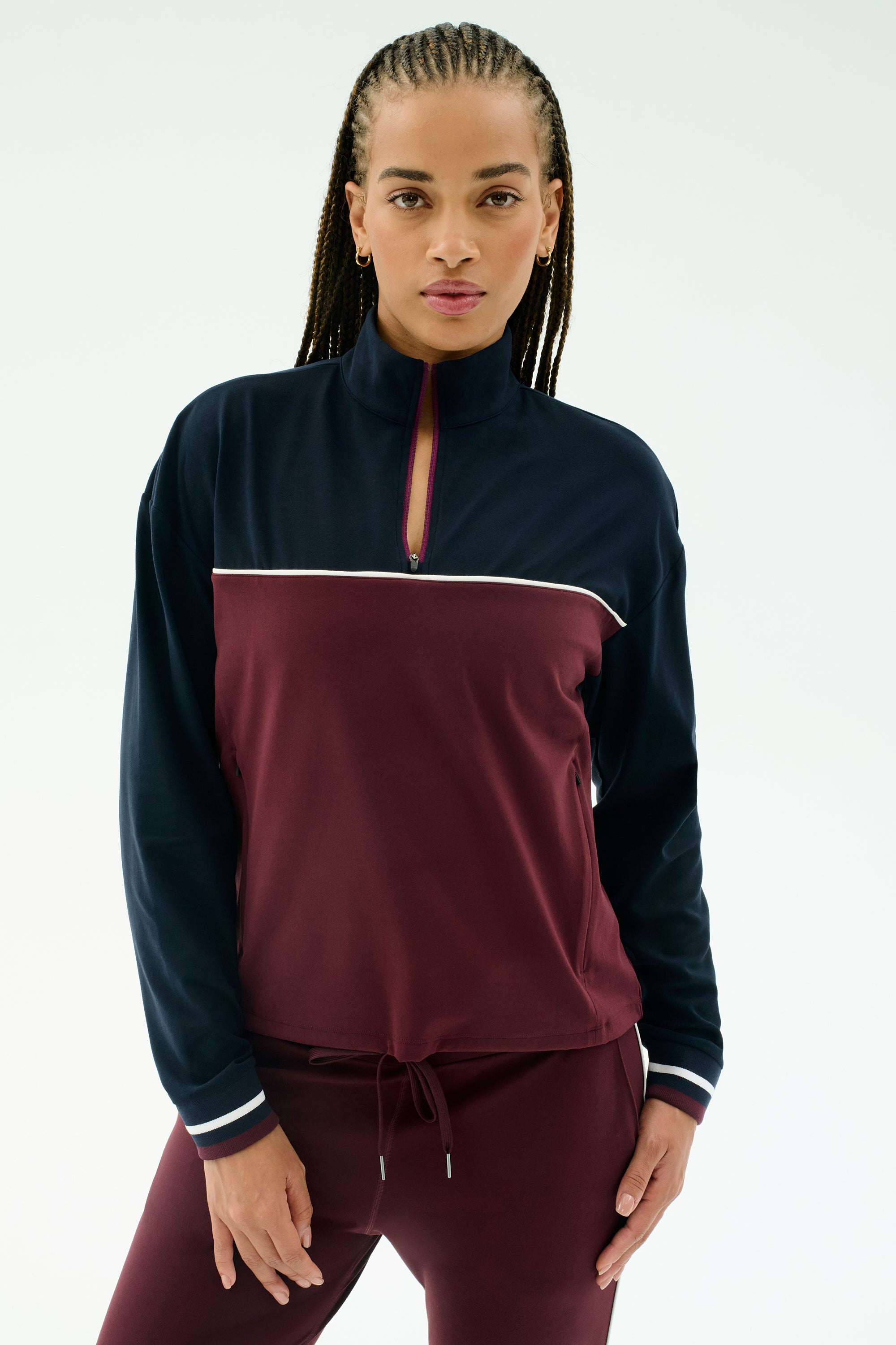 A woman wears the SPLITS59 Millie Airweight Half Zip in Dark Cherry/Indigo with matching pants, made from a nylon-spandex blend, standing against a plain light background.