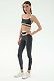 A woman stands against a plain background wearing the SPLITS59 Eva Rigor Double Strap Bra in Graphite/White with matching leggings and white athletic shoes, creating a stylish gym-ready look.