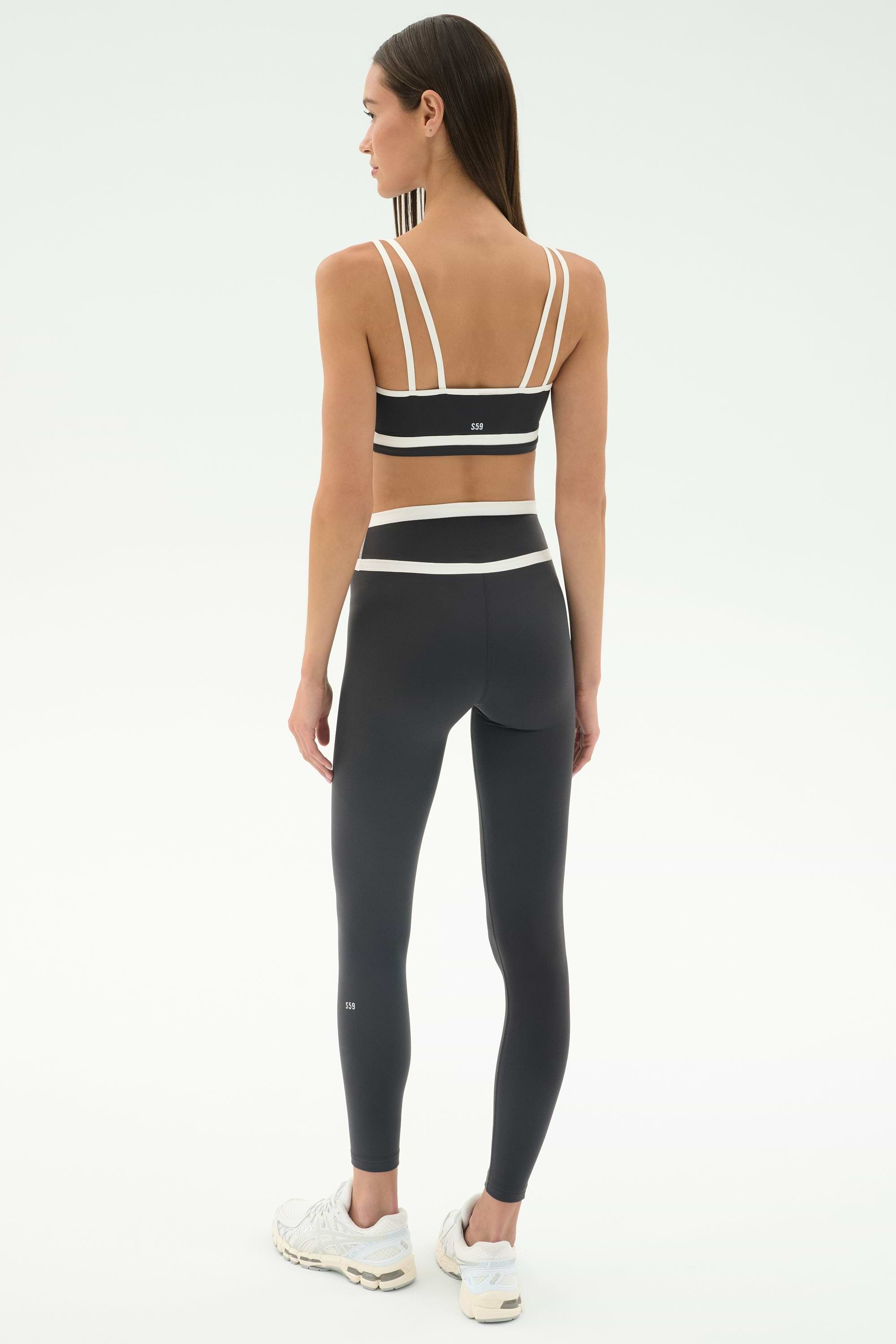 A woman faces away, wearing SPLITS59's Mason High Waist Rigor Full Length leggings in Graphite/White with a matching sports bra and white sneakers, set against a plain light background.