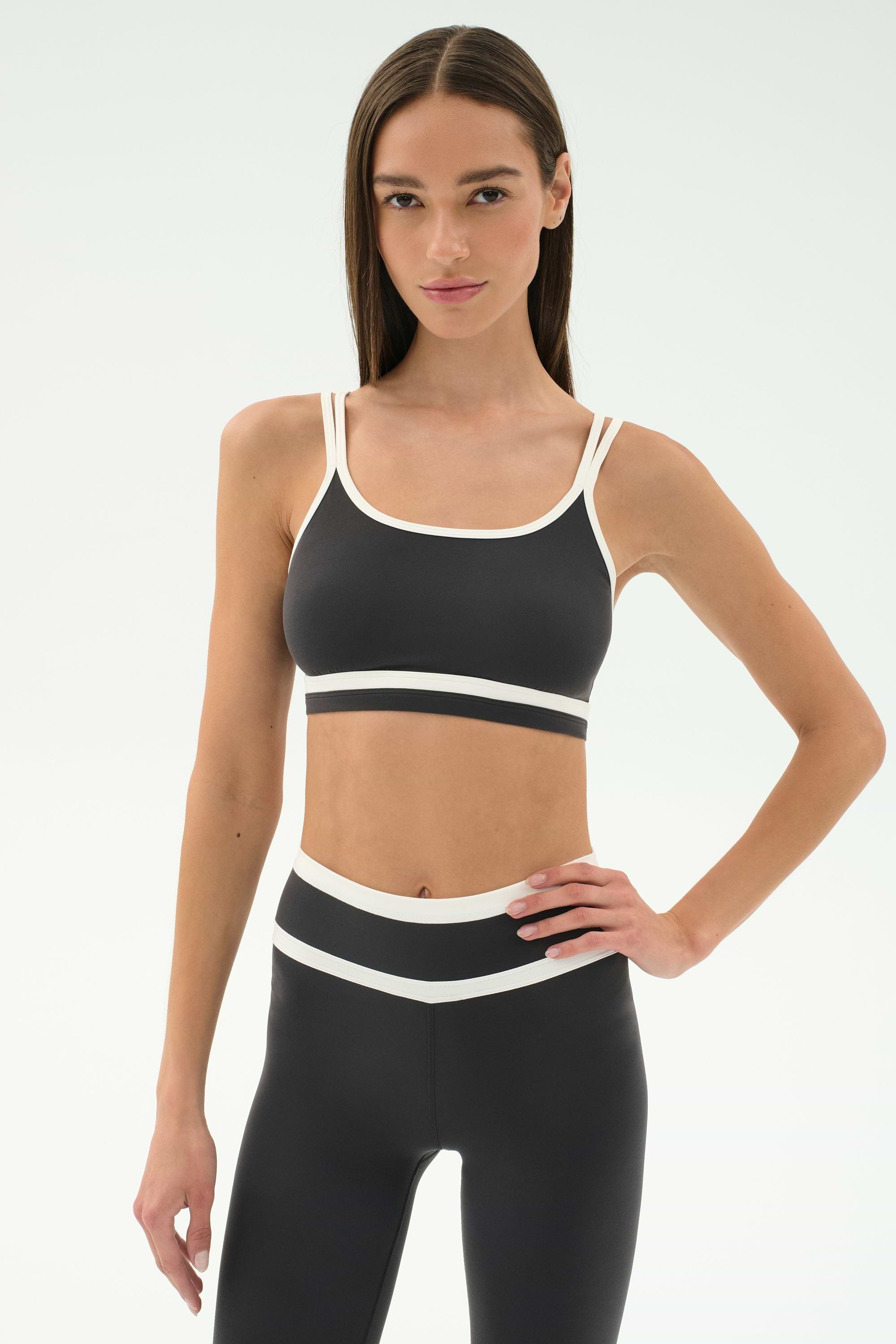 Woman wearing the SPLITS59 Eva Rigor Double Strap Bra in Graphite/White, standing with one hand on her hip against a plain background—ideal activewear for gym workouts.