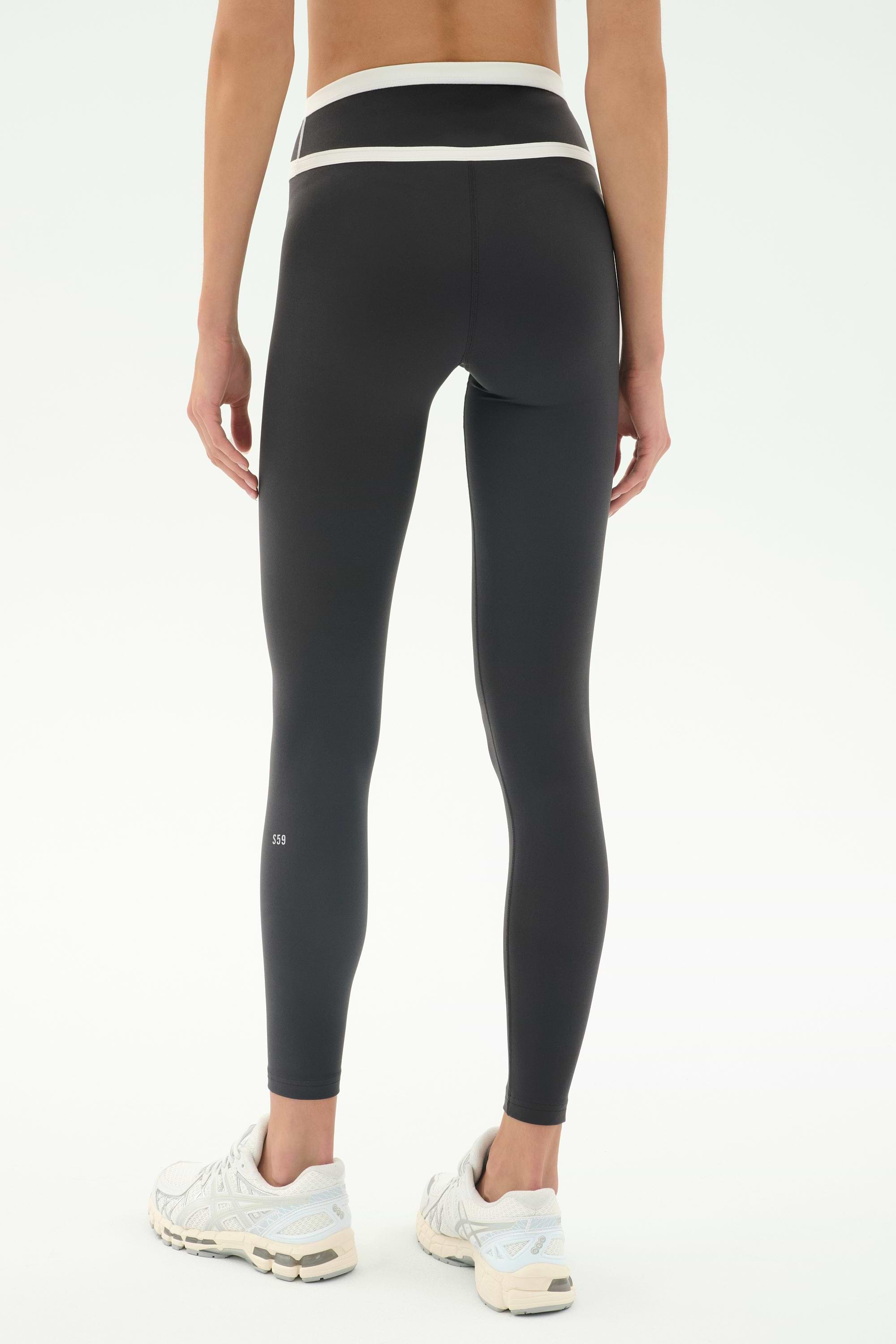 A person wearing SPLITS59 Mason High Waist Rigor Full Length leggings in Graphite/White, paired with white sneakers, stands facing away from the camera against a plain background.