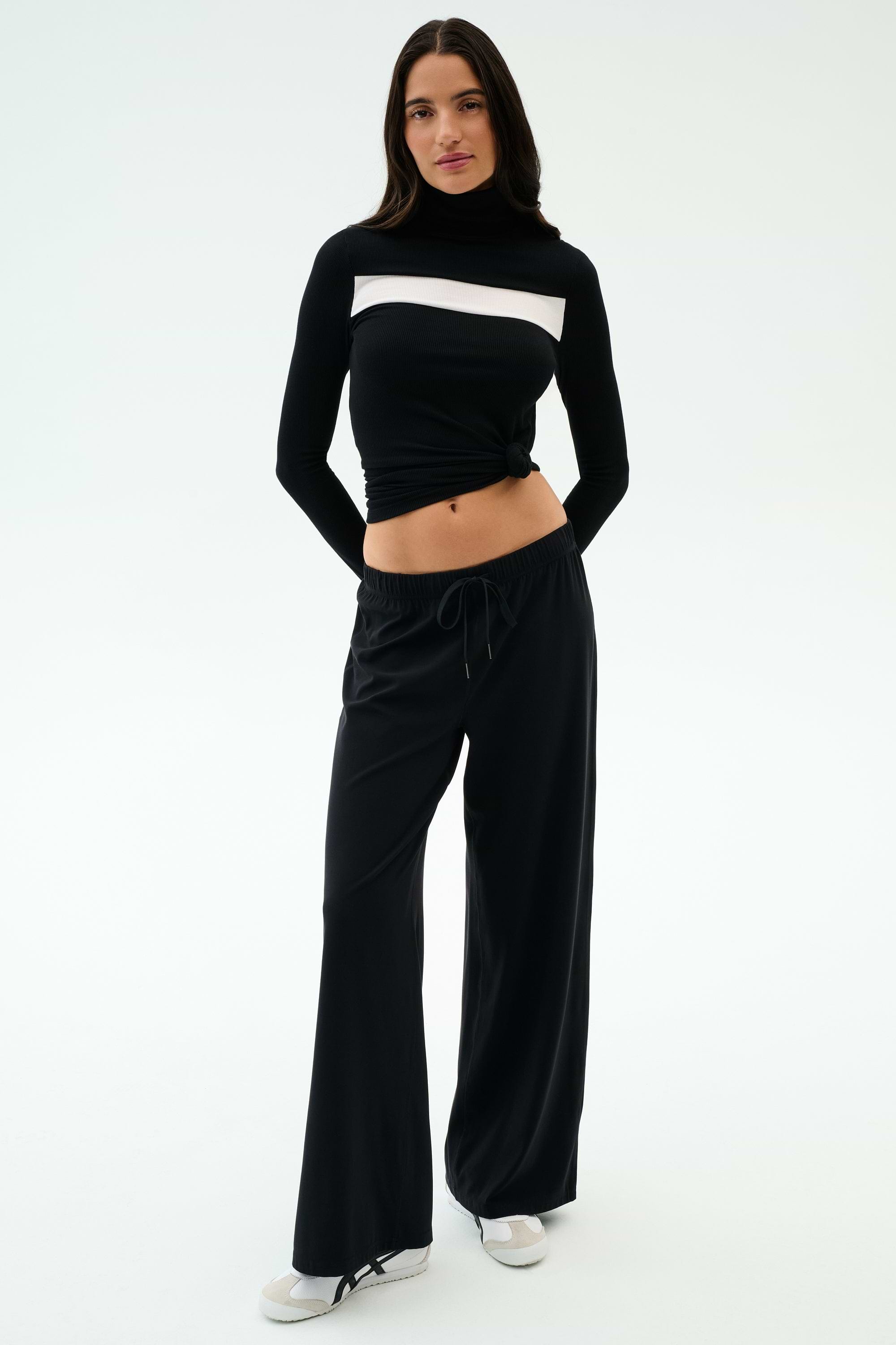 A woman stands facing forward against a plain background wearing SPLITS59's Brynn Airweight Pant in black, paired with a black nylon long-sleeve turtleneck featuring a white horizontal stripe and white shoes.