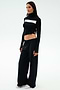 A woman stands against a plain background, wearing the SPLITS59 Brynn Airweight Pant in black, paired with a black and white long-sleeve spandex front-knot top and white sneakers.