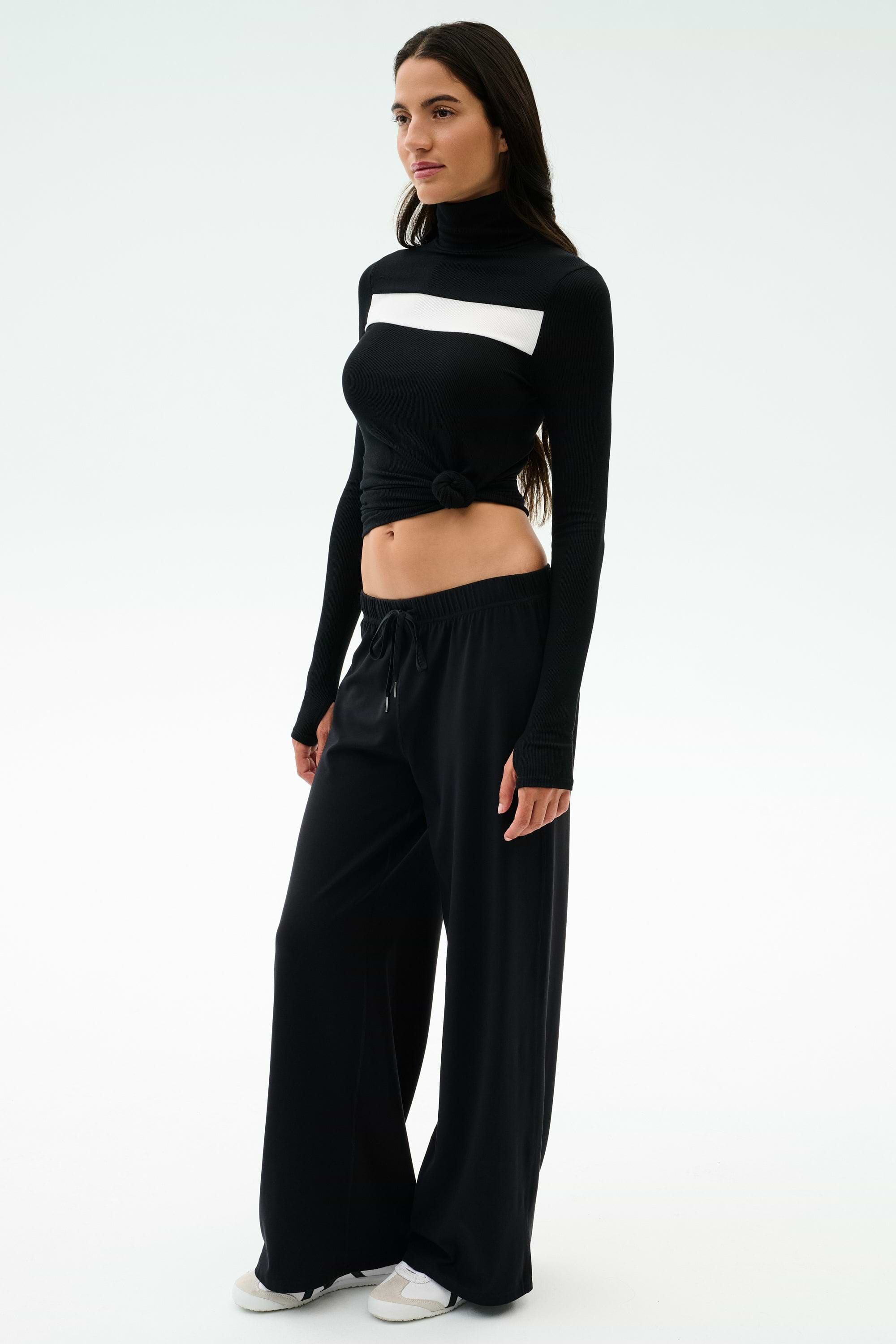 A woman stands against a plain background, wearing the SPLITS59 Brynn Airweight Pant in black, paired with a black and white long-sleeve spandex front-knot top and white sneakers.