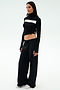 A woman stands against a plain background, wearing the SPLITS59 Brynn Airweight Pant in black, paired with a black and white long-sleeve spandex front-knot top and white sneakers.