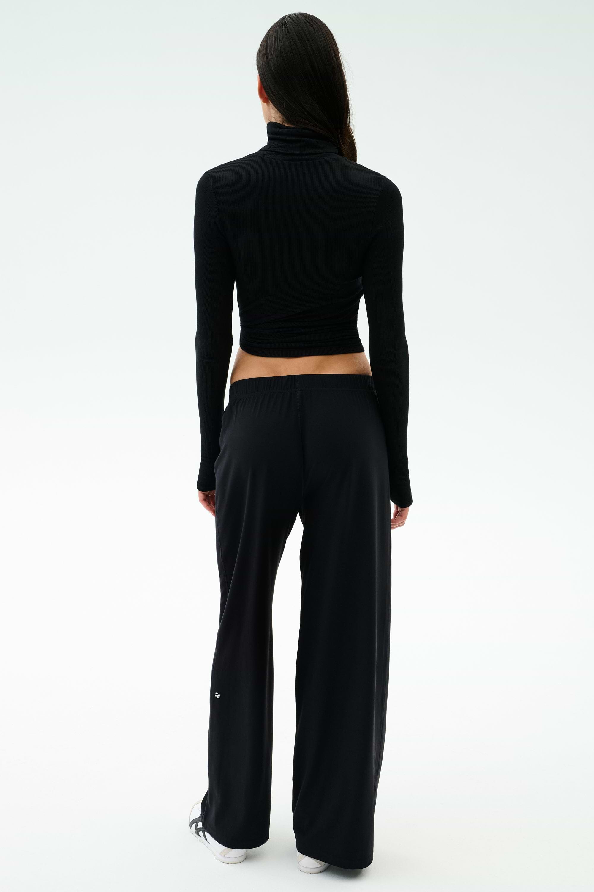 A person faces away, wearing SPLITS59's Brynn Airweight Pant in black, paired with a black spandex long-sleeve turtleneck and white sneakers against a plain light background.