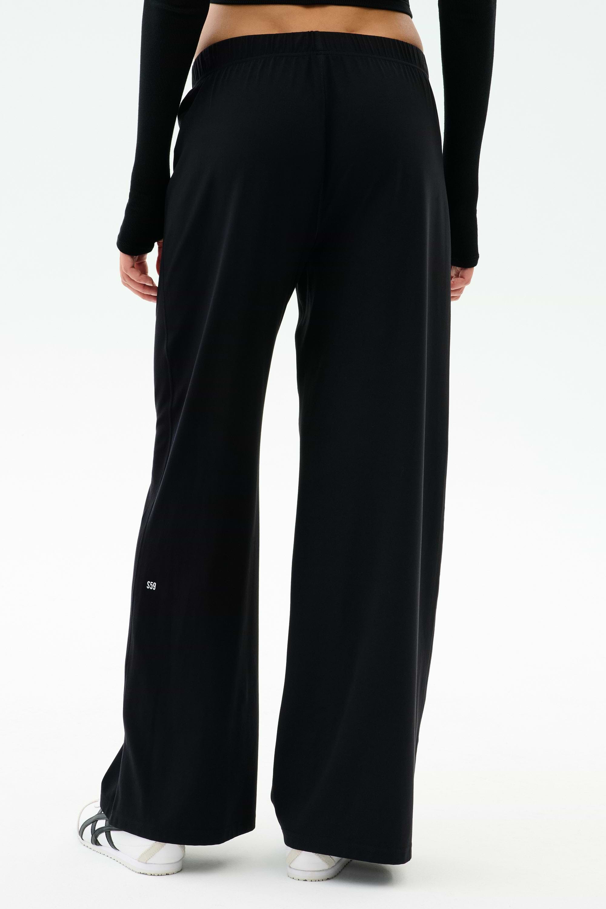 Shown from the back against a plain white background, a person wears SPLITS59 Brynn Airweight Pant in black with a black long-sleeve spandex top and white shoes.