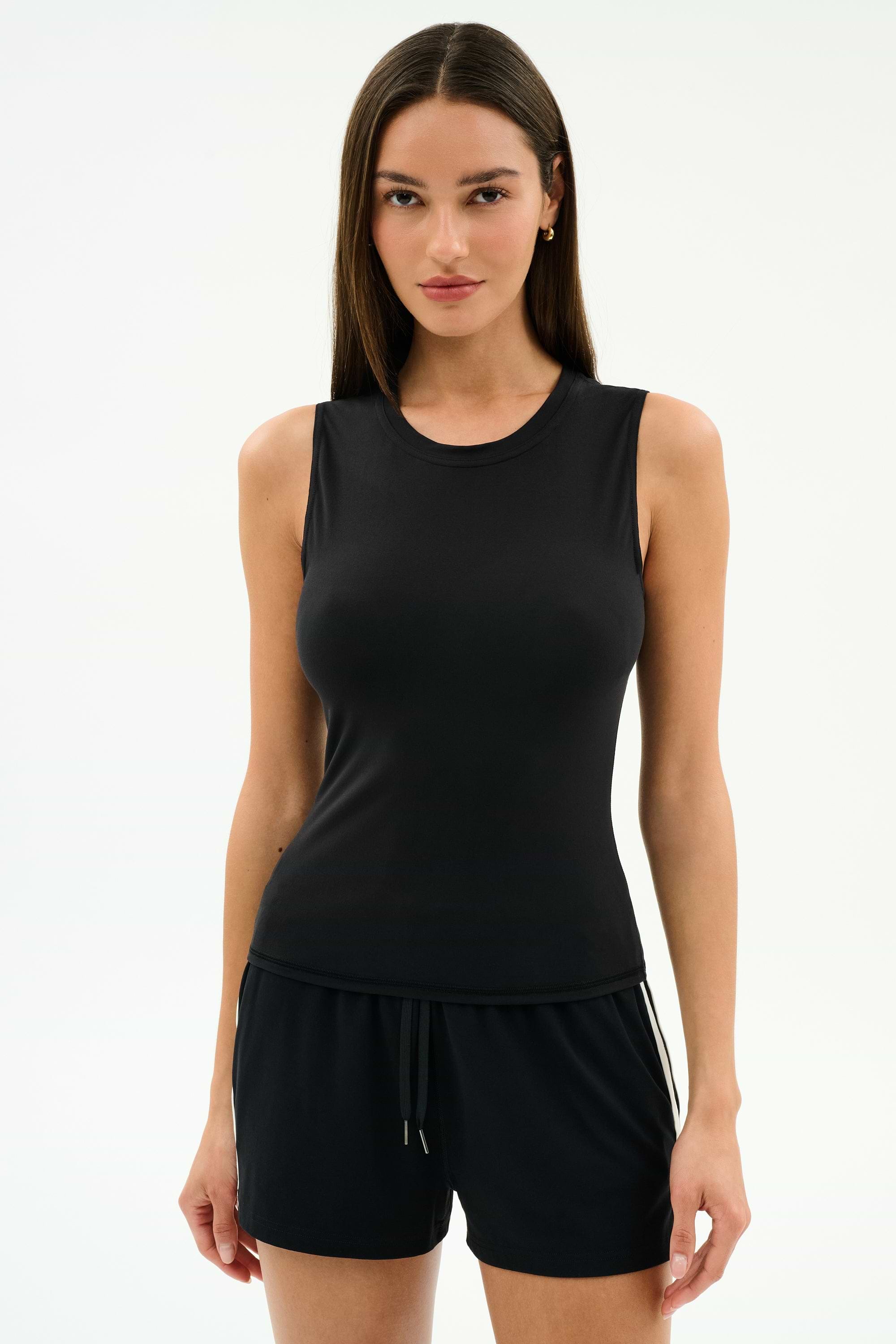 A woman stands against a plain background, wearing the SPLITS59 Carter Airweight Lite Tank in black and black spandex shorts.