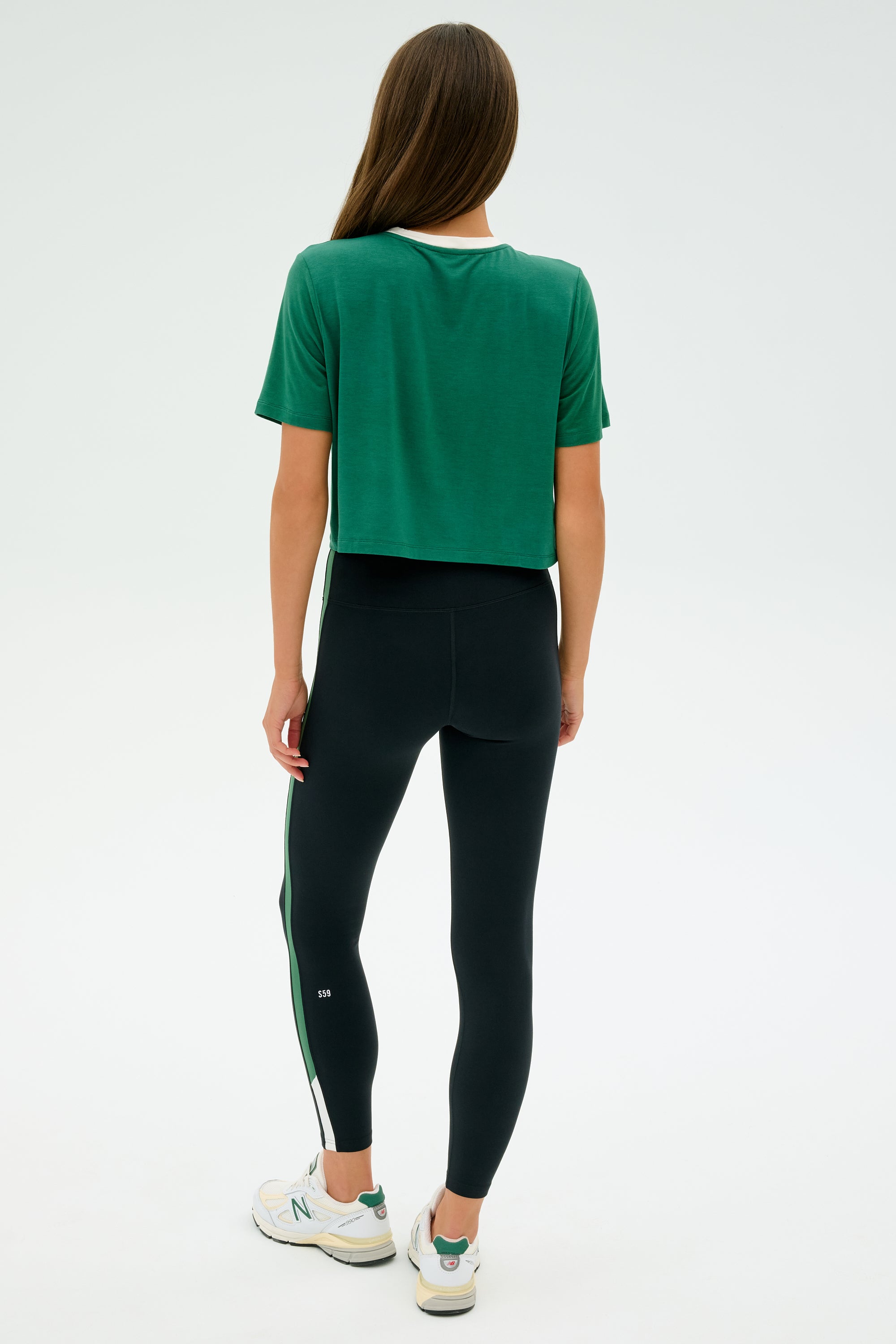 A person wearing the SPLITS59 Djuna Crop Ringer in Arugula/White, black leggings, and white sneakers stands facing away on a plain background.