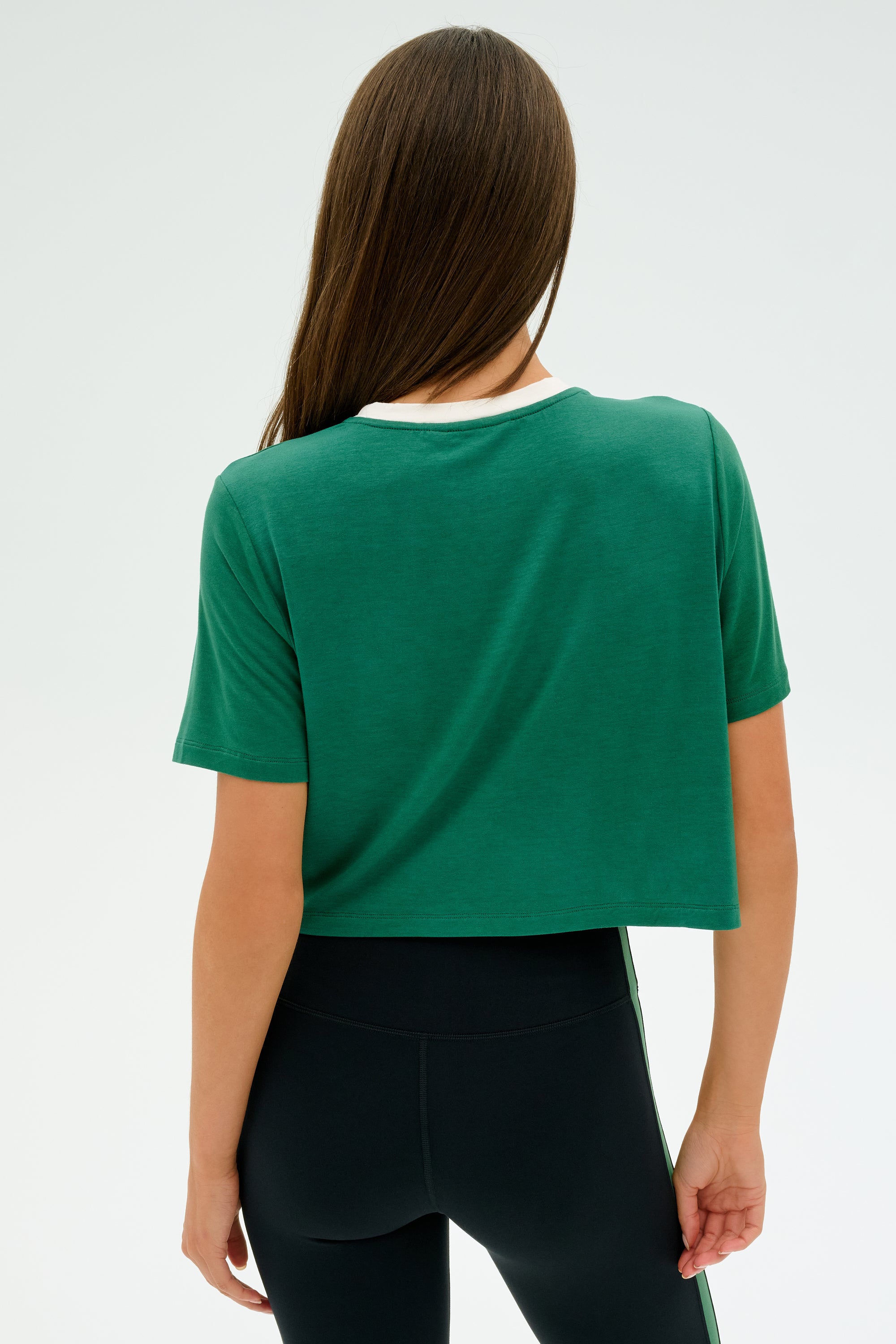 A person with long brown hair is facing away, wearing the SPLITS59 Djuna Crop Ringer in Arugula/White with black leggings. The outfit, made from a viscose and elastane blend, drapes effortlessly against the plain light background.