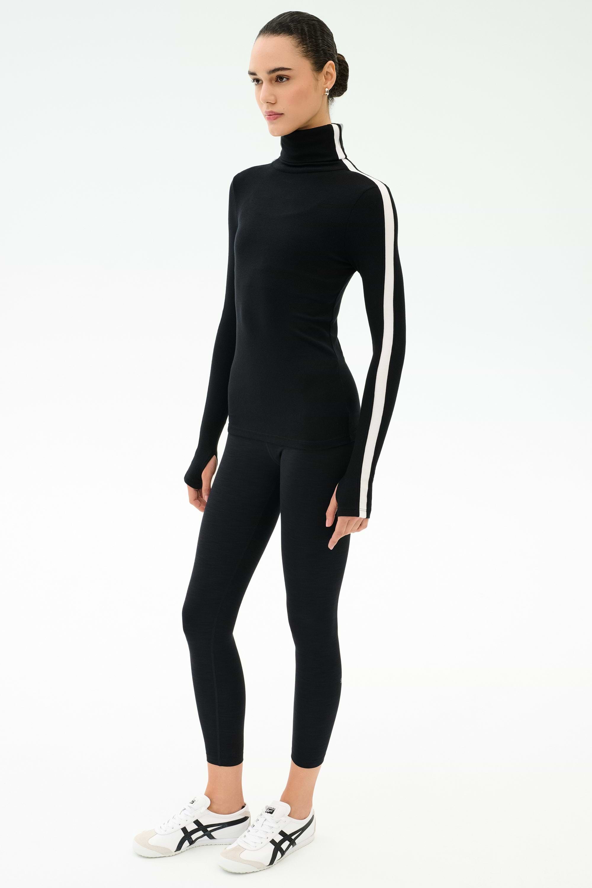 A person wears the SPLITS59 Jackson Rib Full Length Turtleneck in Black/White, paired with black leggings and white sneakers, standing against a plain white background.