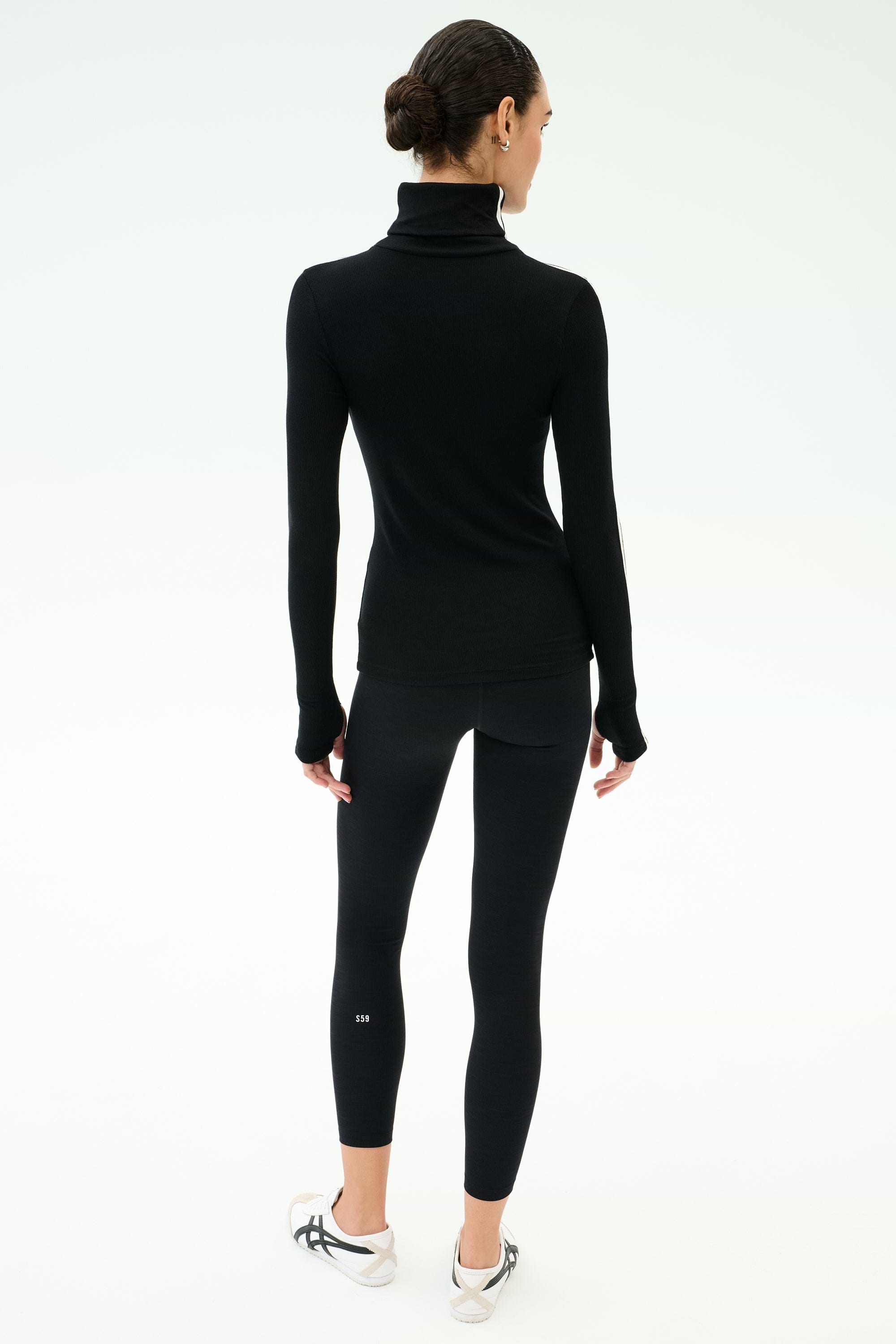A woman faces away against a plain light background, wearing the SPLITS59 Jackson Rib Full Length Turtleneck in Black/White, paired with black leggings and white sneakers.