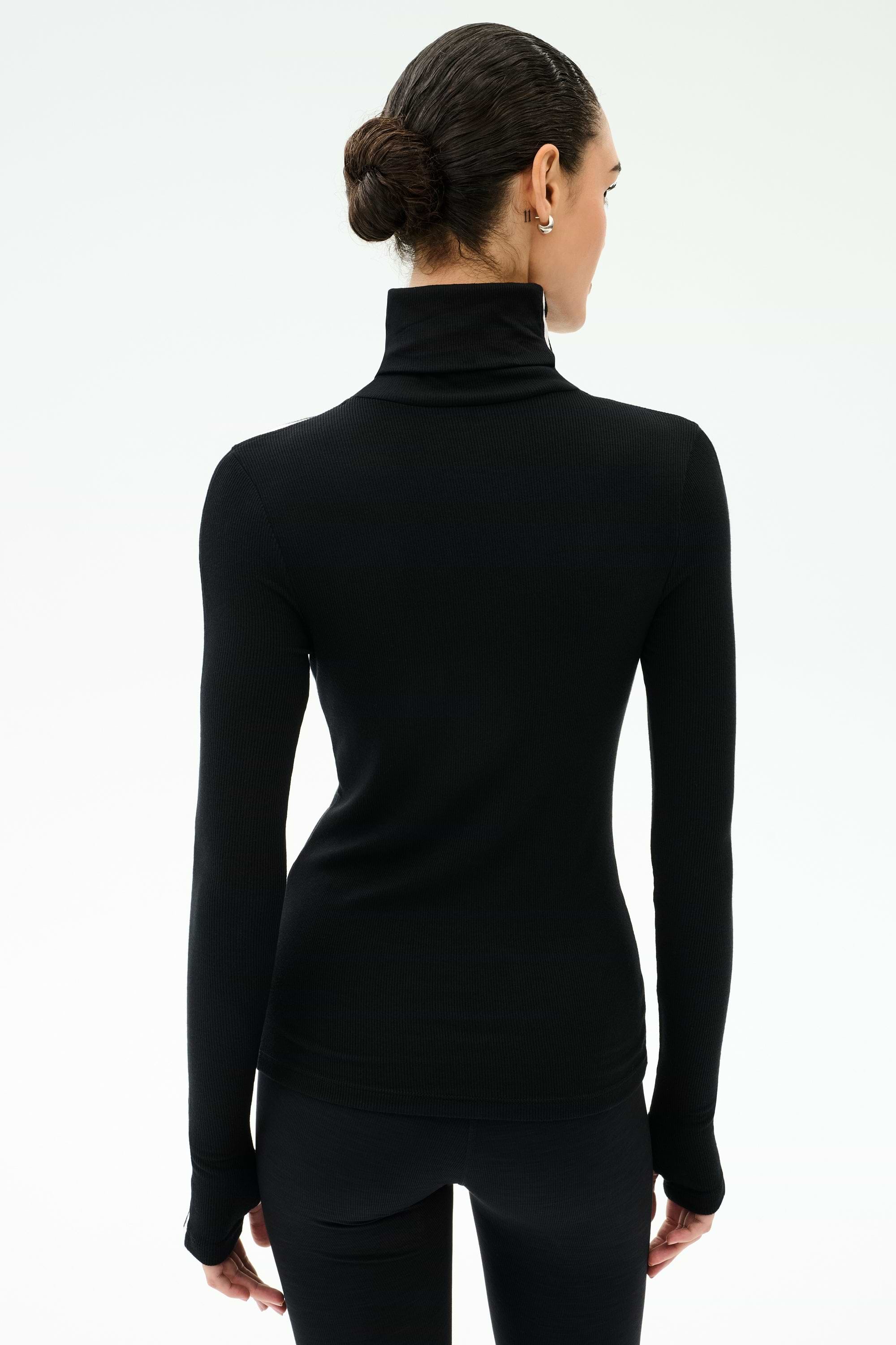A person with dark hair in a low bun wears the SPLITS59 Jackson Rib Full Length Turtleneck - Black/White and black pants, standing against a plain light background, facing away from the camera.