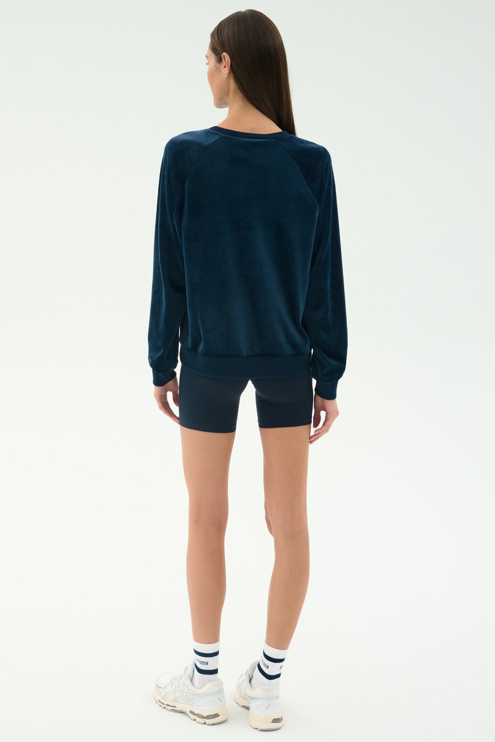 A person stands facing away, wearing the SPLITS59 Joy Velour Crewneck in Indigo, navy shorts, white socks, and white sneakers against a plain light background.