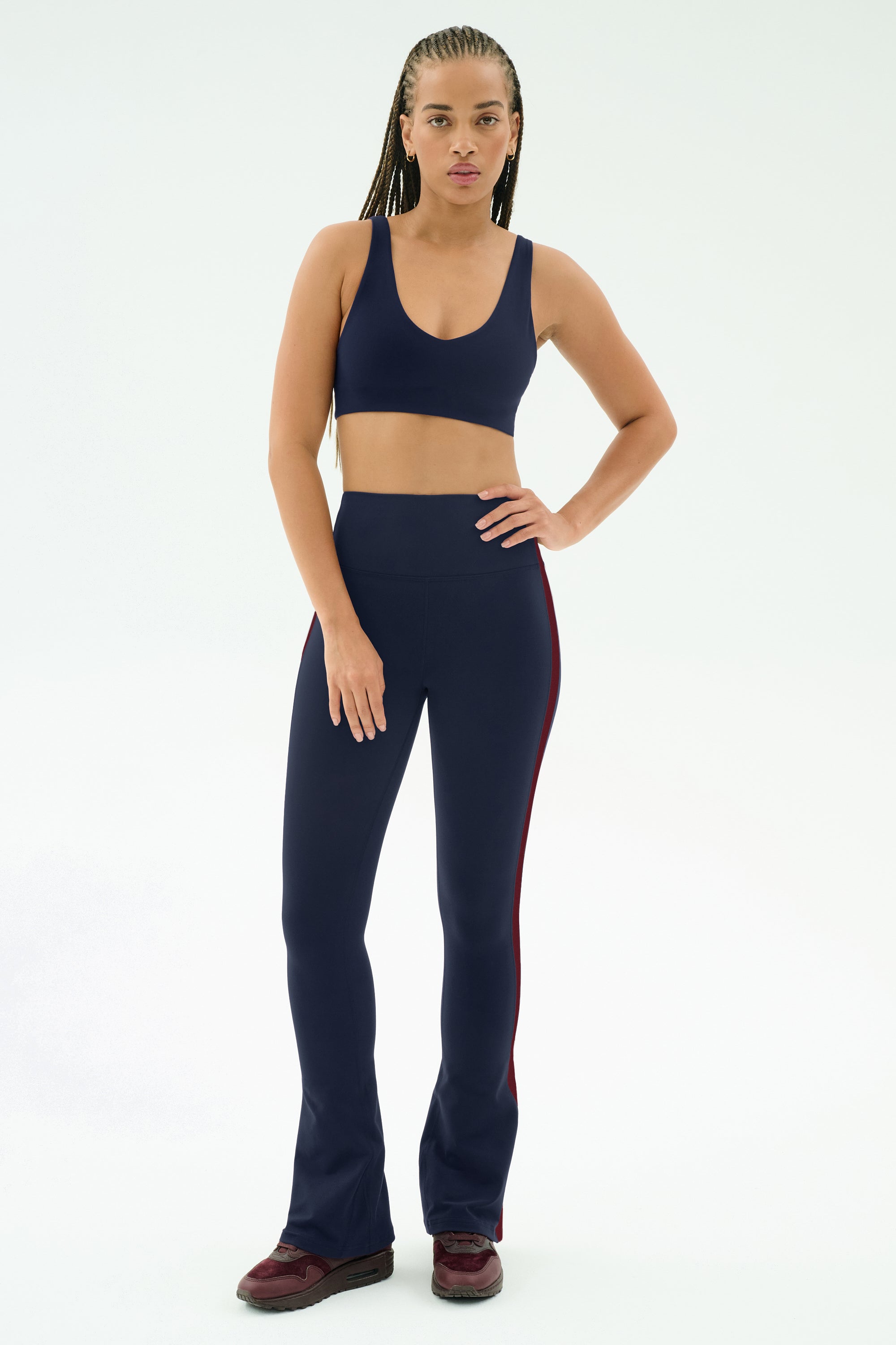 A person faces forward, wearing the SPLITS59 Raquel High Waist Flared Legging in Indigo/Dark Cherry and a navy sports bra, paired with burgundy sneakers against a plain white background.