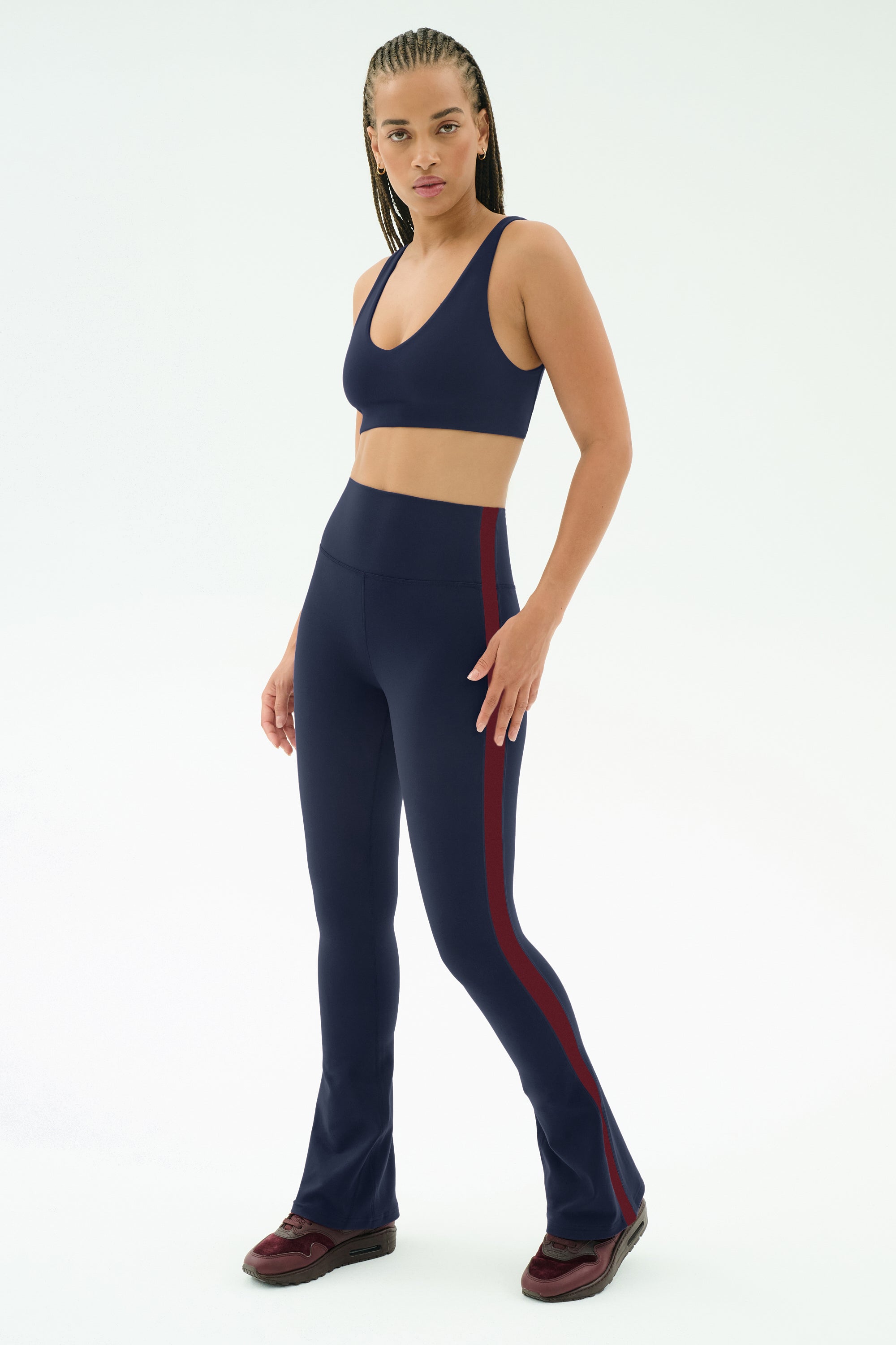 A woman poses in a studio wearing SPLITS59’s Raquel High Waist Flared Legging in Indigo/Dark Cherry and a matching navy sports bra, styled with burgundy sneakers.