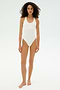 A woman with curly hair stands barefoot against a plain white background, wearing the SPLITS59 Suki Airweight Bodysuit in white, a smooth nylon sleeveless one-piece.