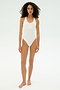 A woman with curly hair stands barefoot against a plain white background, wearing the SPLITS59 Suki Airweight Bodysuit in white, a smooth nylon sleeveless one-piece.