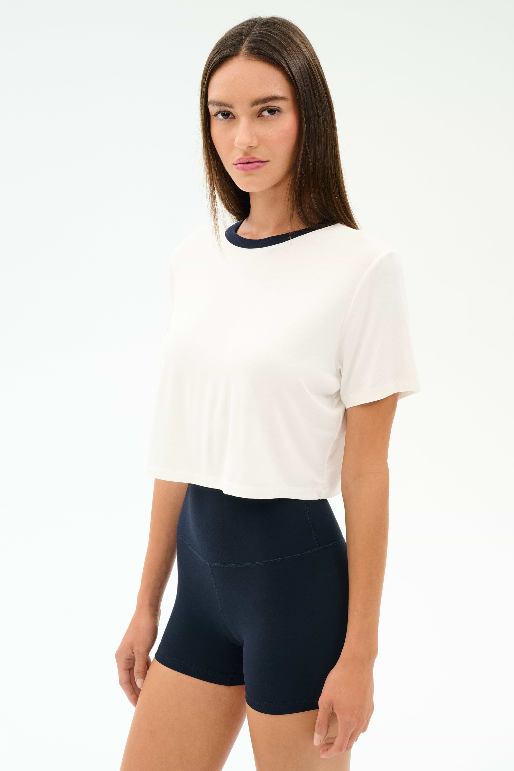 A woman stands against a plain background wearing the SPLITS59 Djuna Crop Ringer in White/Indigo with high-waisted black shorts—an ideal look for gym workouts or CrossFit sessions.