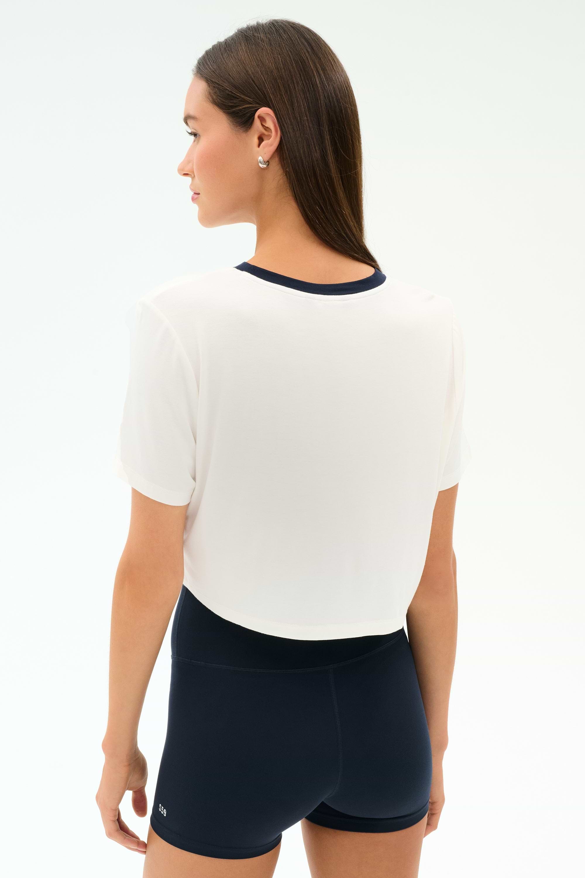 A woman faces away against a plain light background, ready for the gym in the SPLITS59 Djuna Crop Ringer - White/Indigo and black high-waisted shorts.