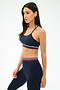 A woman wears the SPLITS59 Cali Active Rib Bra in Heather Indigo with matching leggings featuring red, white, and green striped bands, standing against a plain white background.