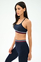 A woman wears the SPLITS59 Cali Active Rib Bra in Heather Indigo with matching leggings featuring red, white, and green striped bands, standing against a plain white background.