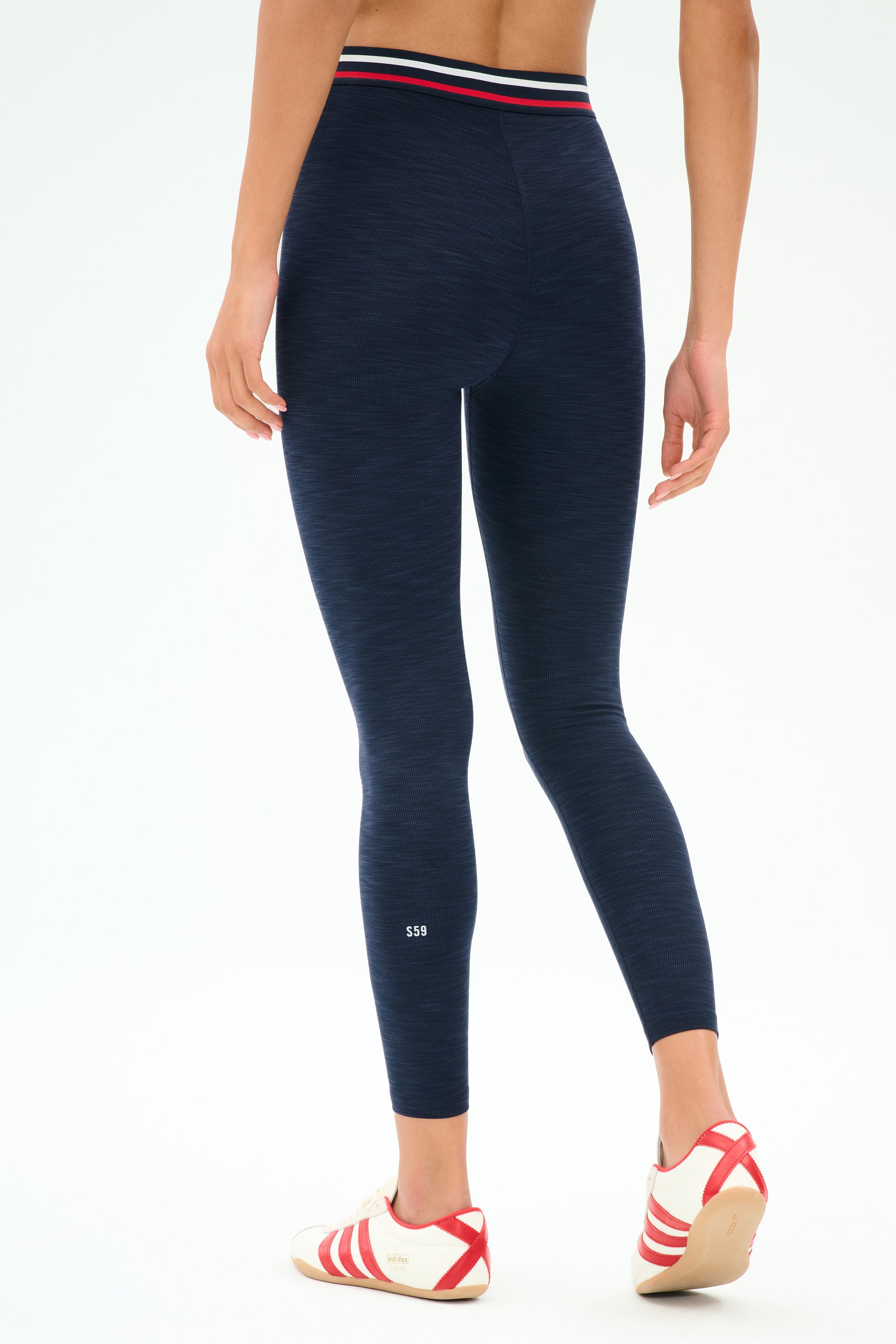 A person is shown from the back wearing SPLITS59's Bailey High Waist Active Rib 7/8 leggings in Heather Indigo, featuring a striped waistband, paired with white sneakers with red stripes—ideal for gym workouts.