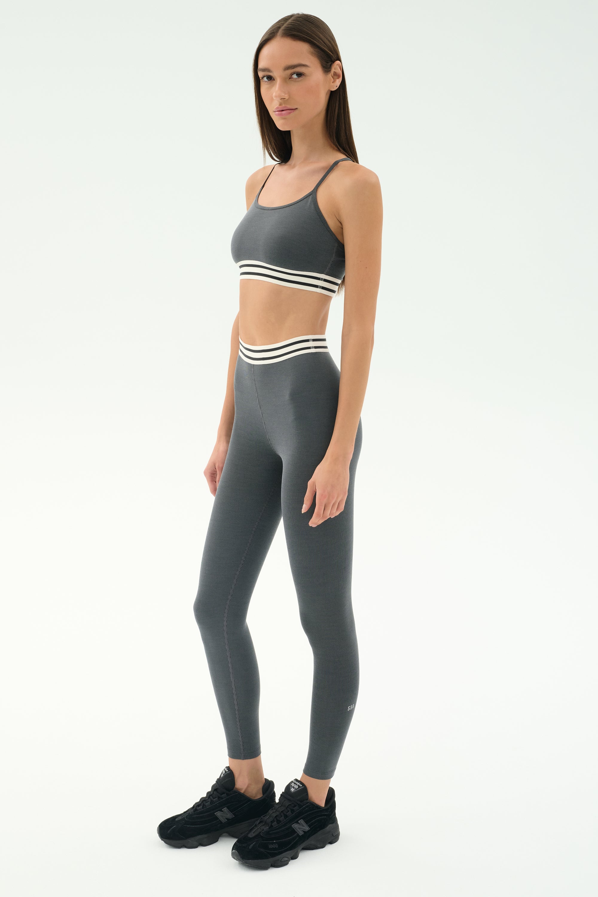 A woman stands against a plain background wearing the SPLITS59 Cali Active Rib Bra in Heather Grey with matching leggings featuring white striped bands, and black sneakers.