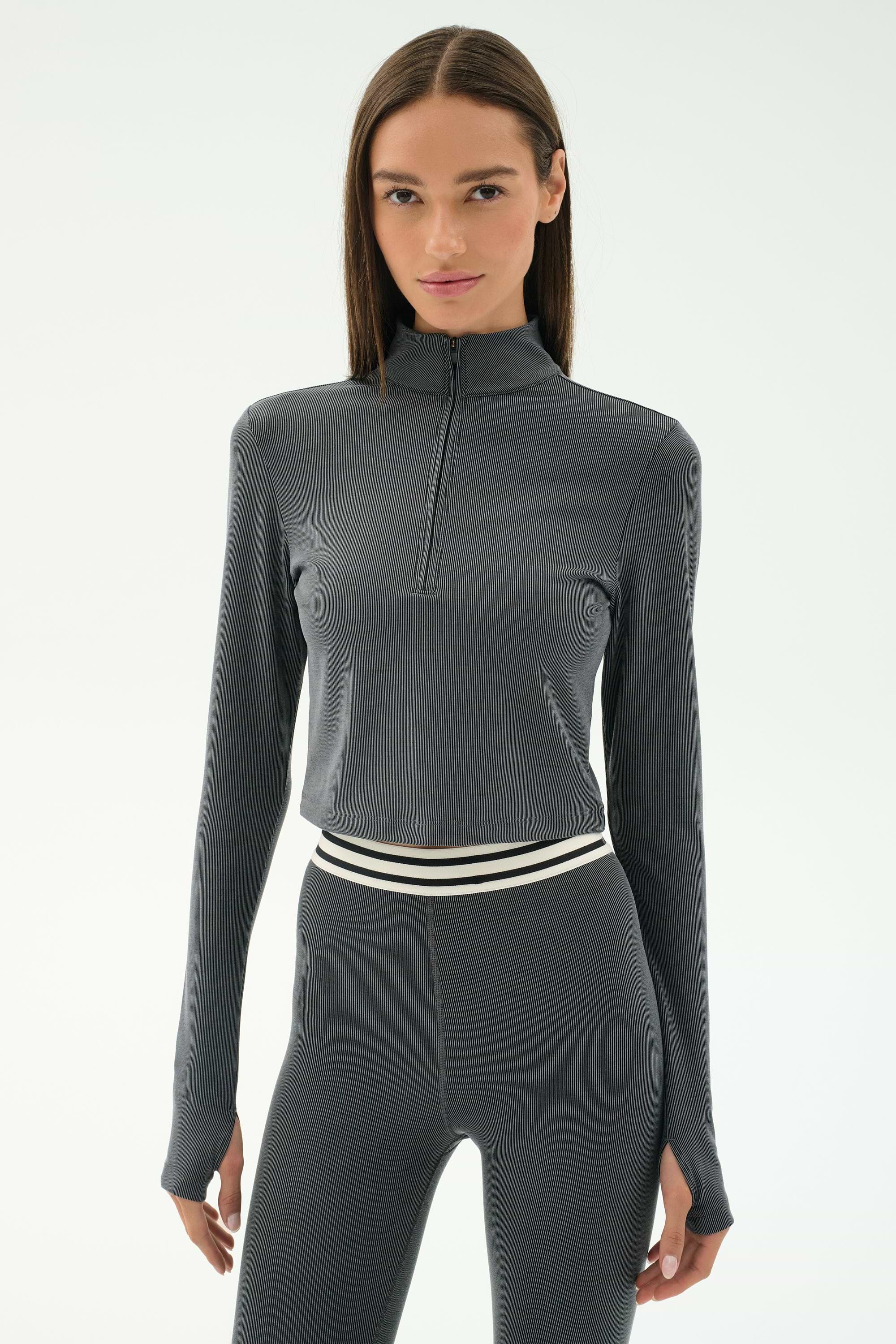 A woman wears the SPLITS59 Stevie Active Rib Half Zip in Heather Grey with matching high-waisted leggings featuring a striped elastic waistband, perfect for gym workouts, standing against a white background.