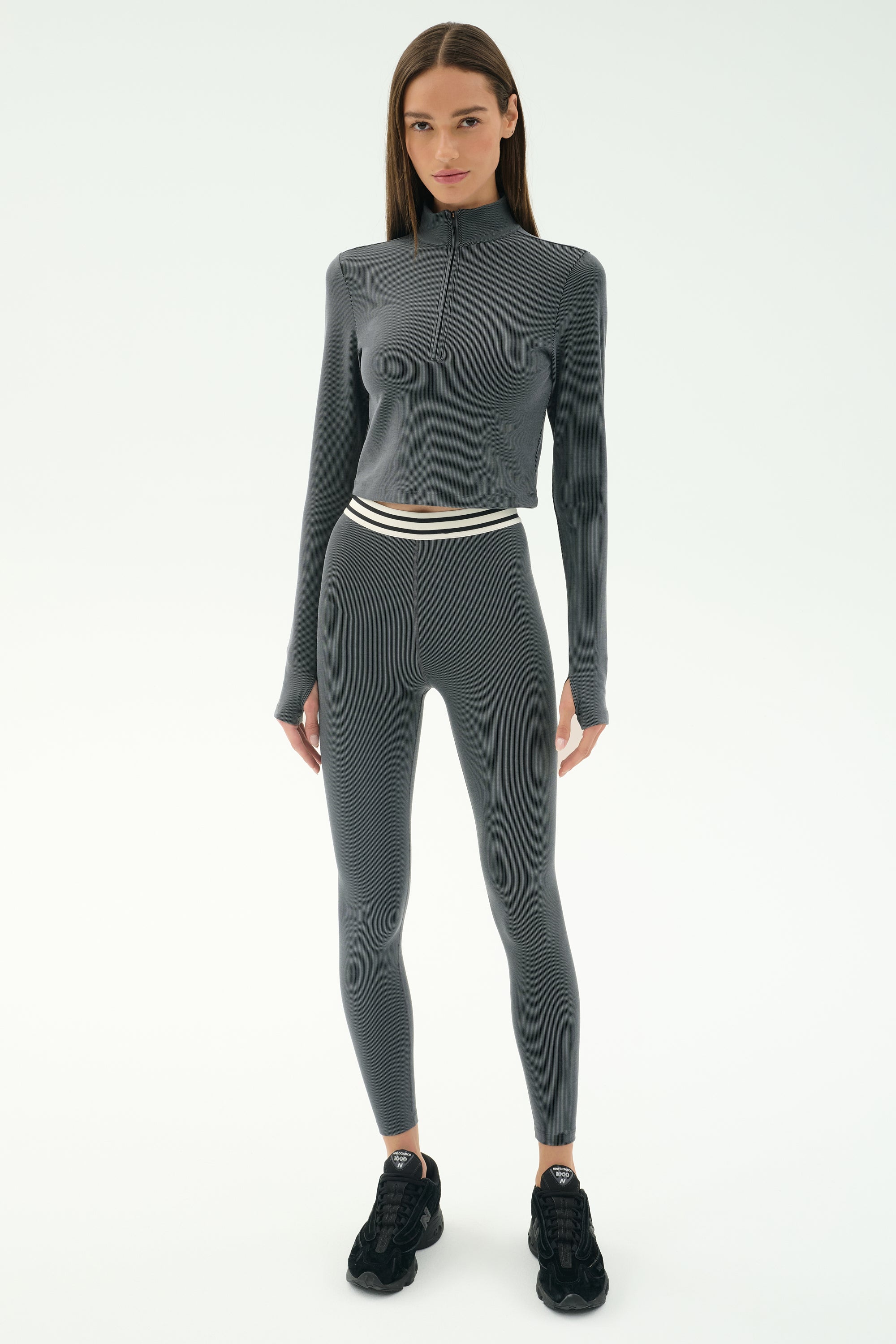 A woman stands against a white background wearing the SPLITS59 Bailey Active Rib High Waist 7/8 leggings in heather grey, a fitted long-sleeve athletic set with subtle stripes and a white-striped waistband, paired with black sneakers.