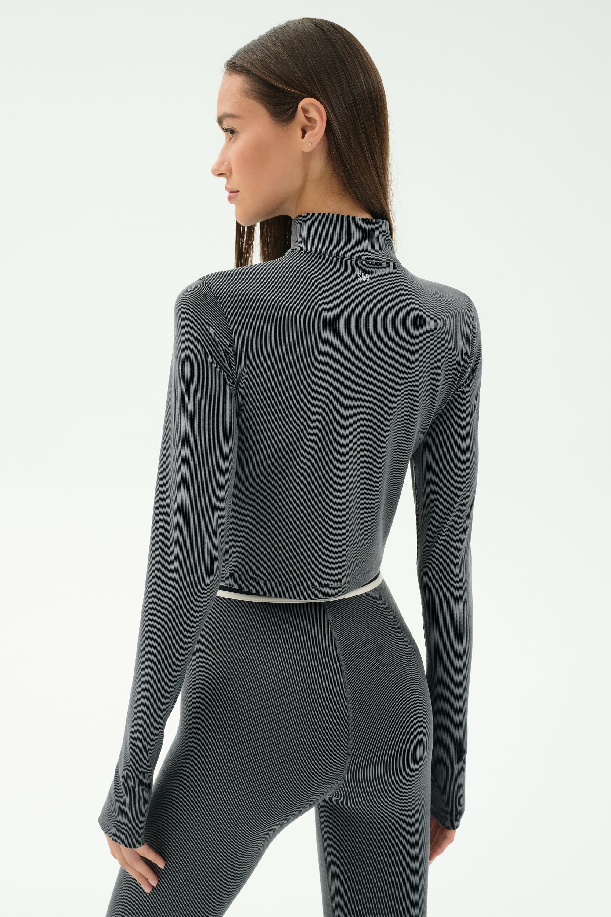 A woman with long straight hair is seen from behind wearing the SPLITS59 Stevie Active Rib Half Zip in Heather Grey with matching leggings, against a plain light background.