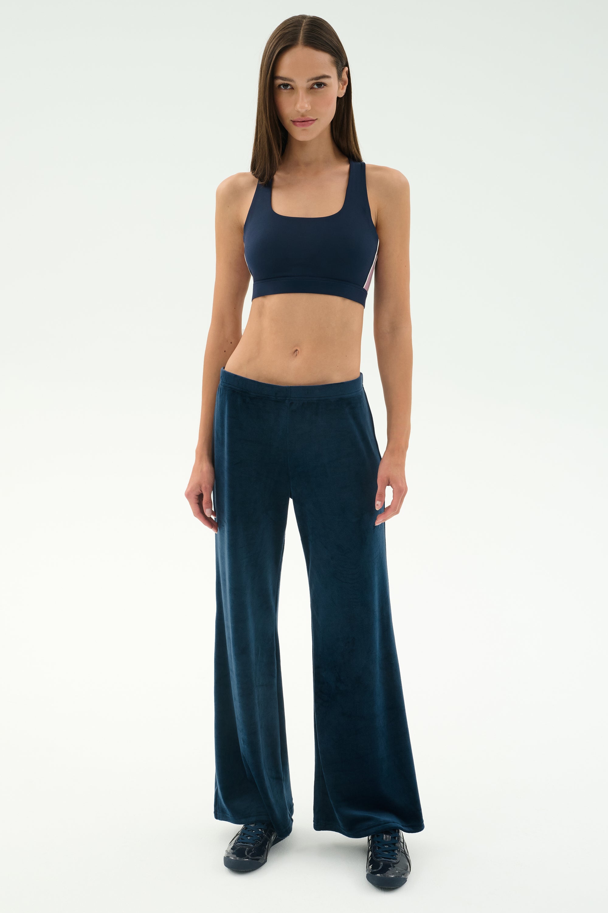 A woman stands facing forward in SPLITS59's Joy Velour Pant - Indigo, paired with a navy sports bra and dark sneakers against a plain light background.