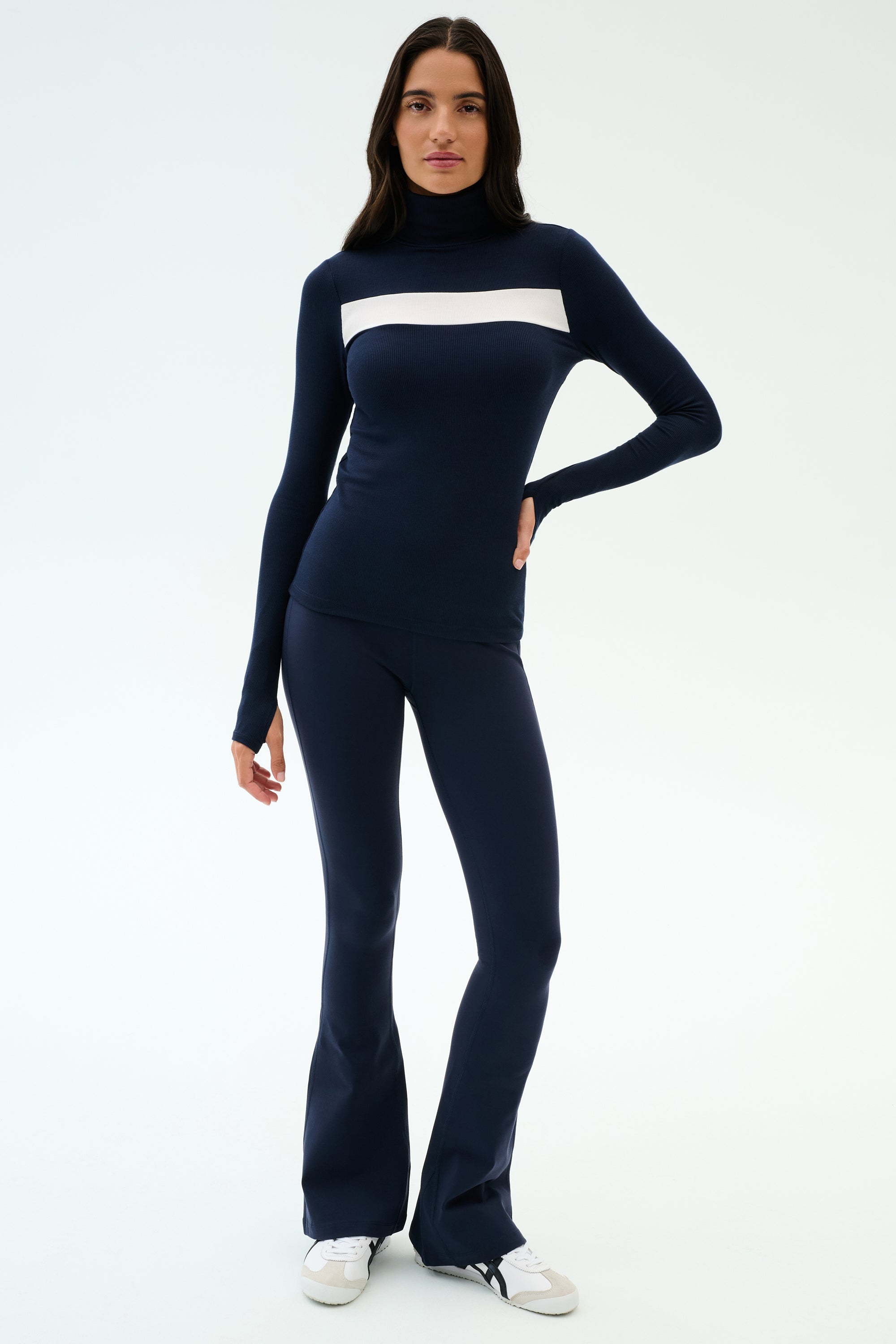 A woman wears the SPLITS59 Lennon Rib Turtleneck in Indigo/White with navy flared pants and white sneakers, standing against a plain light background.