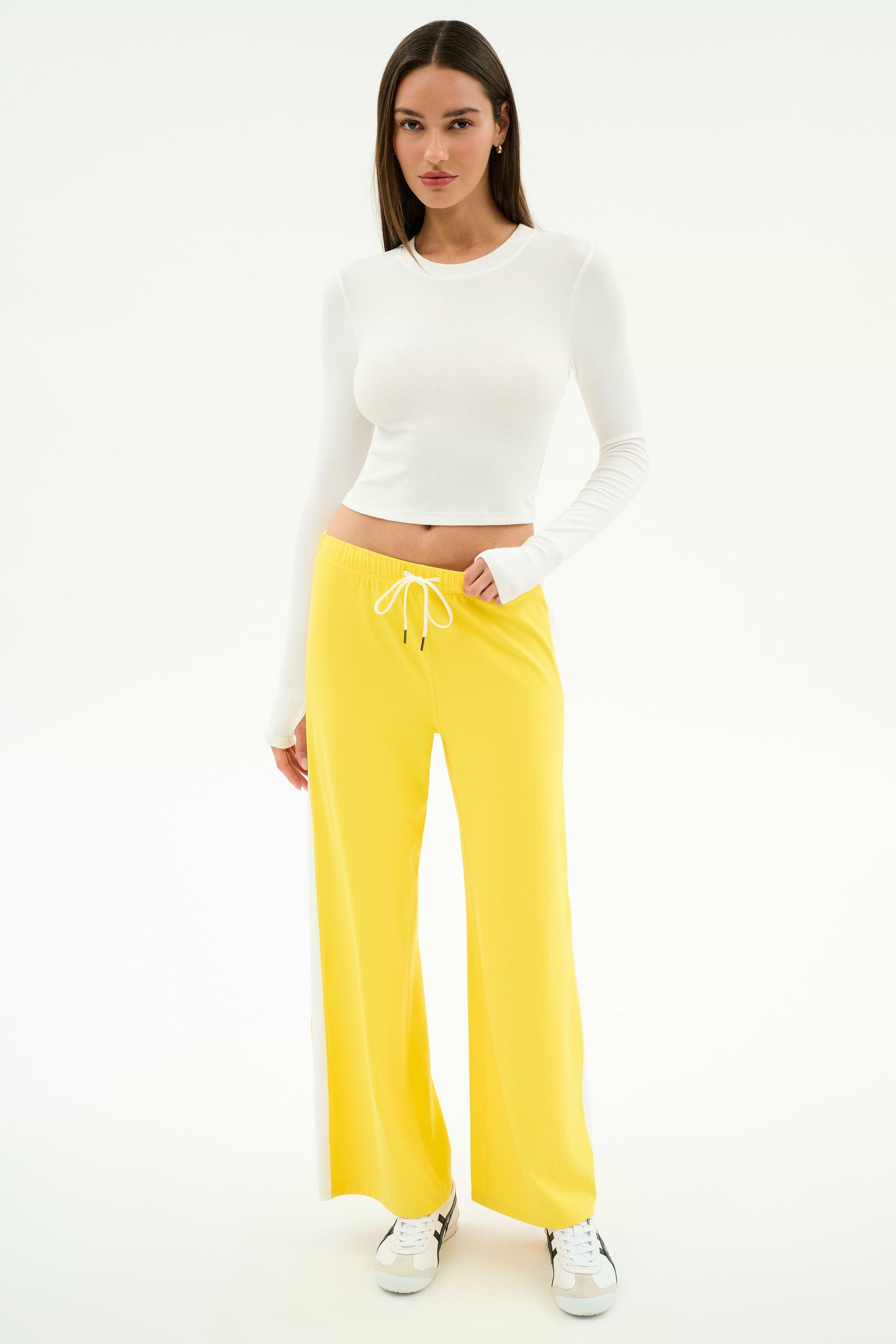 A woman poses against a white background wearing SPLITS59’s Willow Airweight Wide Leg pants in Bright Yellow/White, paired with a long-sleeve white spandex crop top and white sneakers.