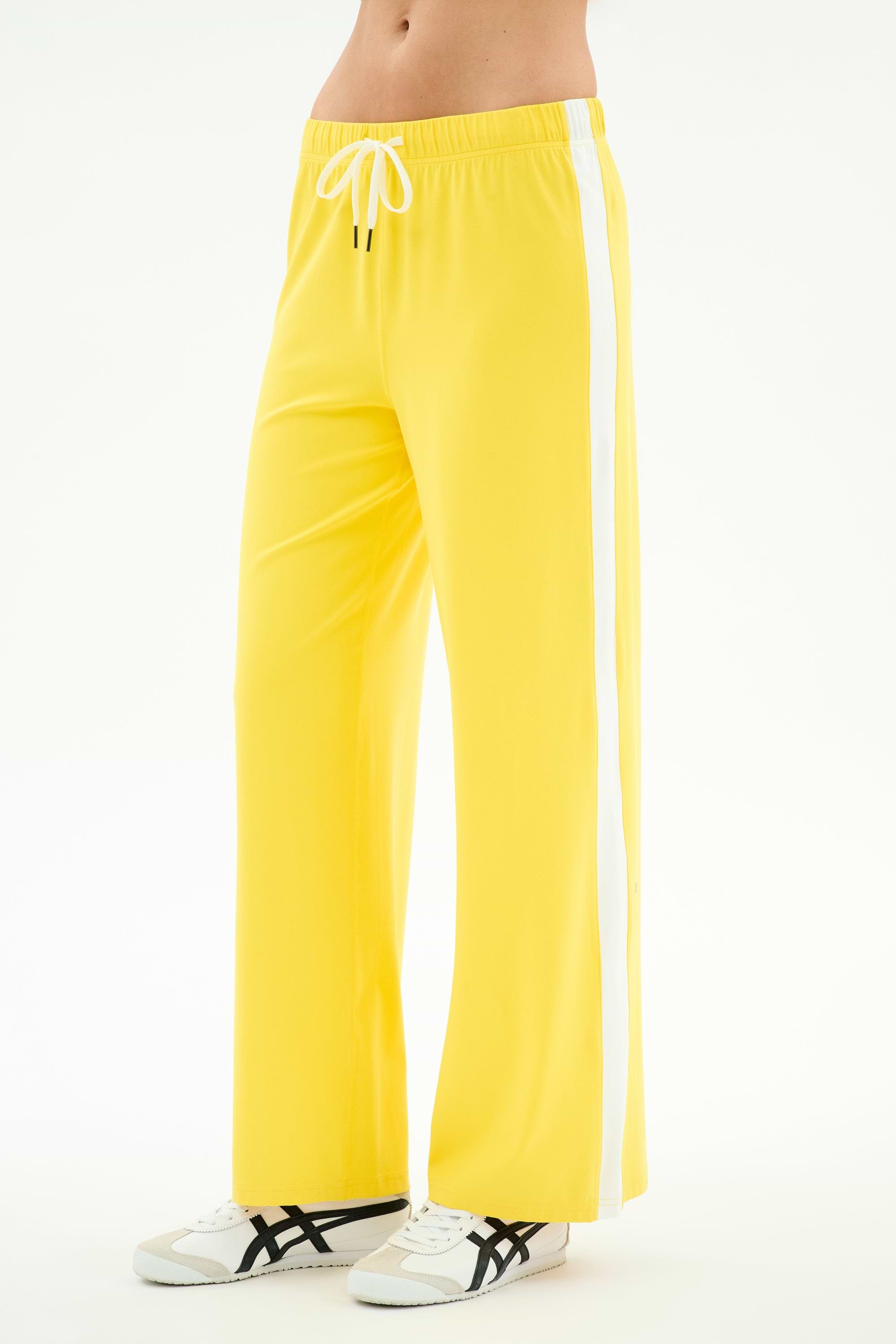 A person wears SPLITS59's Willow Airweight Wide Leg pants in bright yellow/white with white sneakers featuring black accents, standing against a white background.