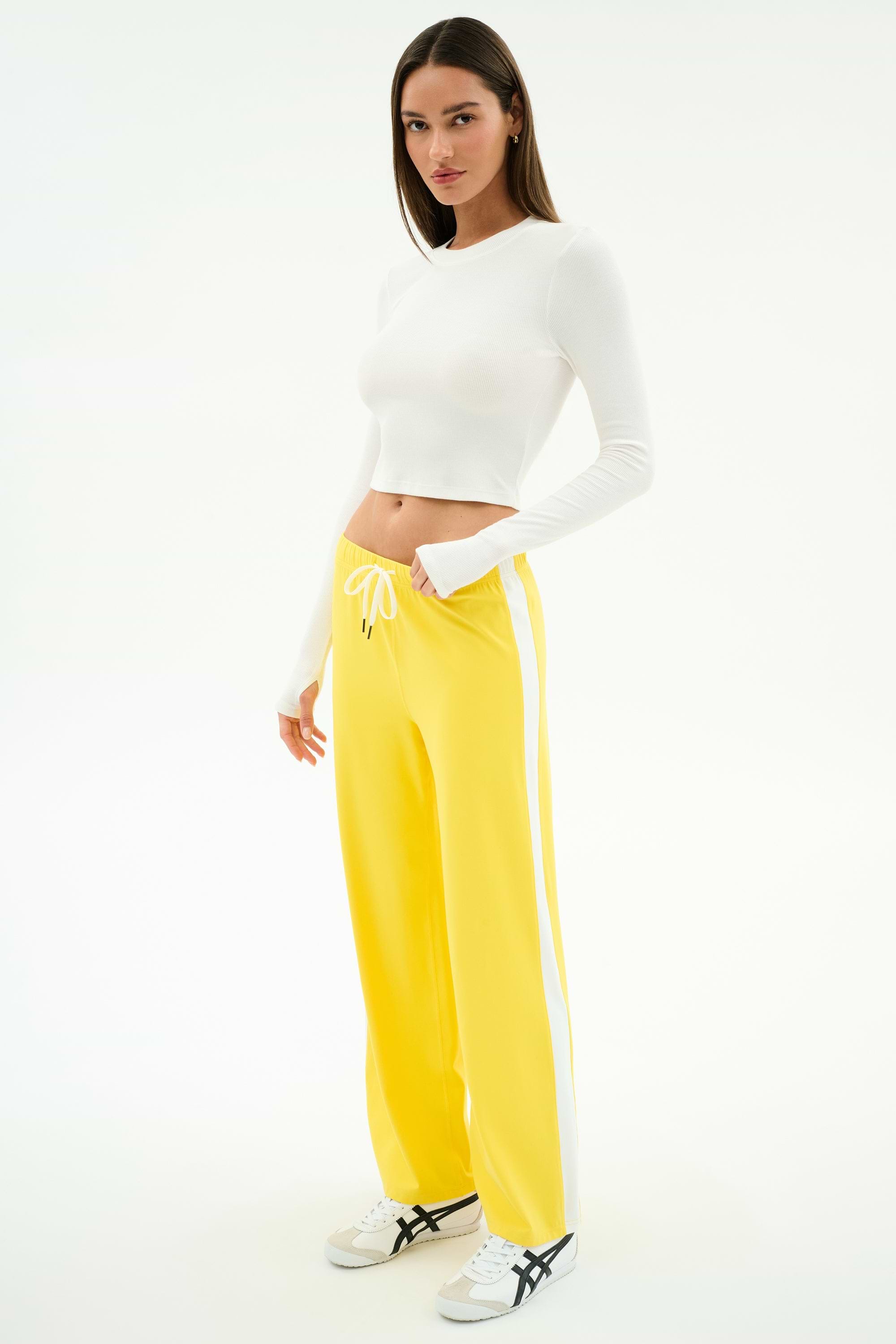 A woman stands against a white background wearing a white spandex long-sleeve crop top, SPLITS59 Willow Airweight Wide Leg pants in Bright Yellow/White, and white sneakers.