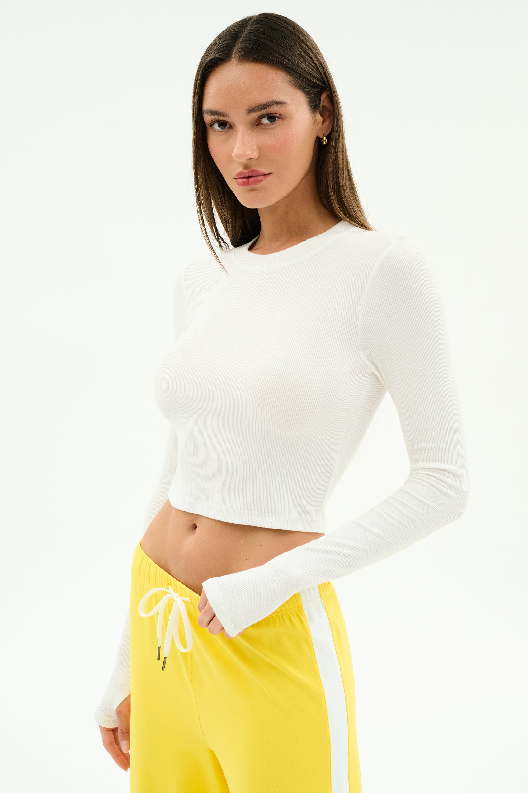 A woman wears the SPLITS59 Louise Rib Long Sleeve Crop in white, paired with yellow drawstring pants, standing against a plain white background.