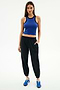 A woman stands against a white background wearing SPLITS59's Megan Woven Nylon Jogger w/ Piping in Black/Creme, paired with a sleeveless blue spandex top and blue sneakers, with one hand in her pocket.