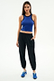 A woman stands against a white background wearing SPLITS59's Megan Woven Nylon Jogger w/ Piping in Black/Creme, paired with a sleeveless blue spandex top and blue sneakers, with one hand in her pocket.