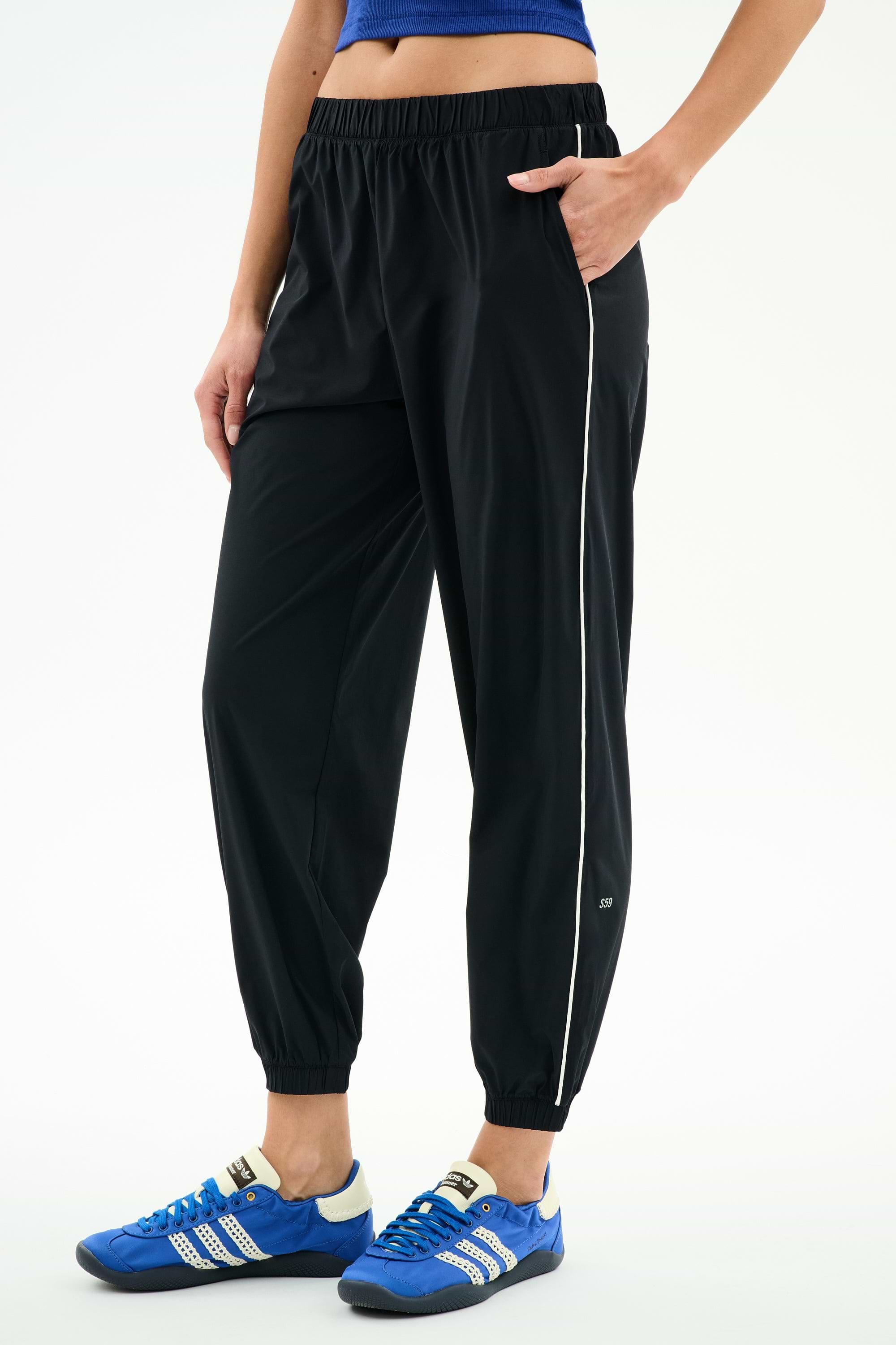 A person stands against a white background wearing SPLITS59 Megan Woven Nylon Jogger w/ Piping - Black/Creme (85% nylon/spandex), featuring a white side stripe, paired with a blue crop top and blue athletic shoes, with one hand in their pocket.