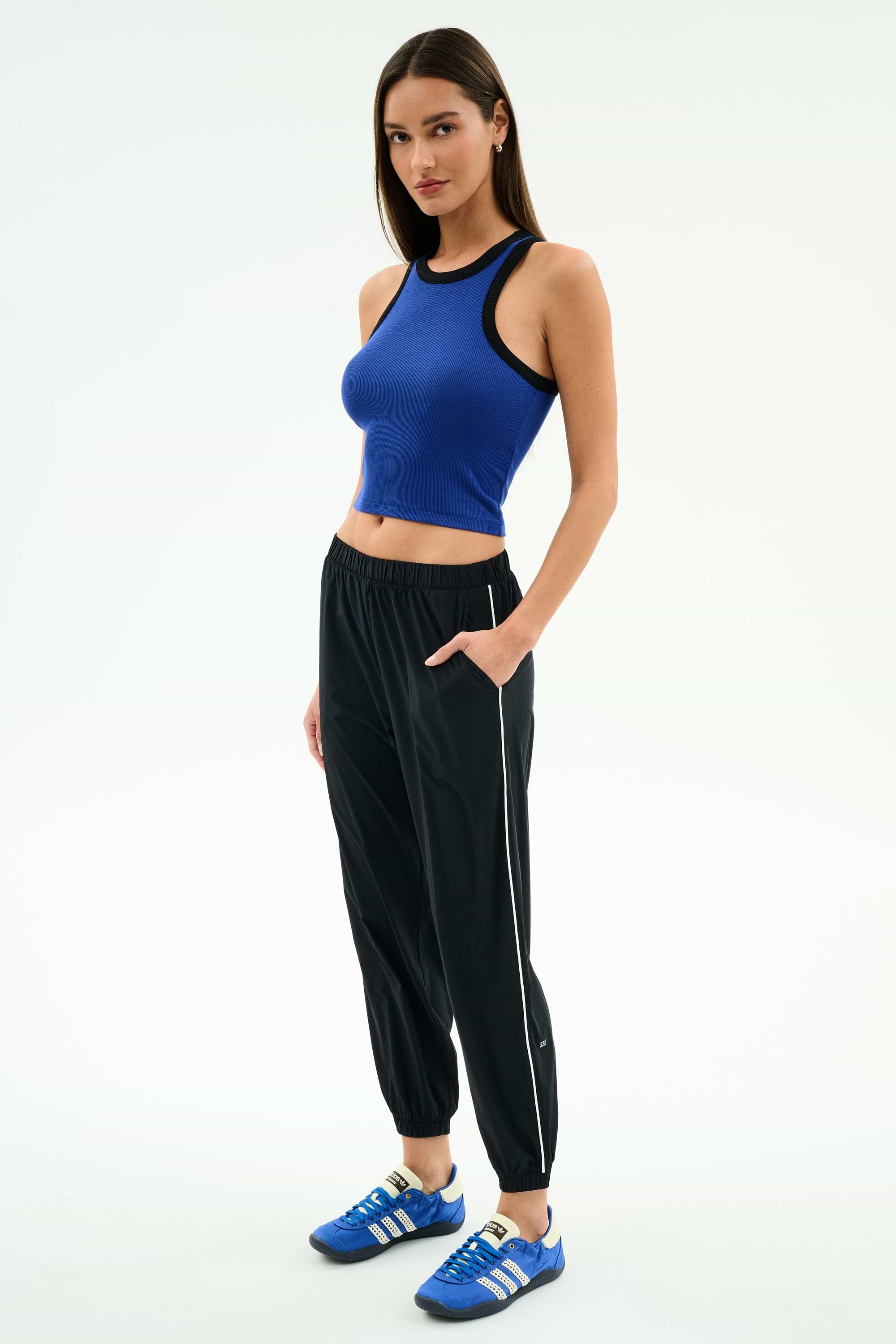 A woman stands against a plain background, wearing a blue sleeveless crop top and SPLITS59 Megan Woven Nylon Jogger w/ Piping in Black/Creme, paired with blue Adidas sneakers.