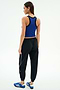 A woman faces away, wearing SPLITS59’s Megan Woven Nylon Jogger w/ Piping in Black/Creme with a blue sleeveless top and blue athletic shoes, crafted from stretchy spandex and 85% nylon, on a plain white background.