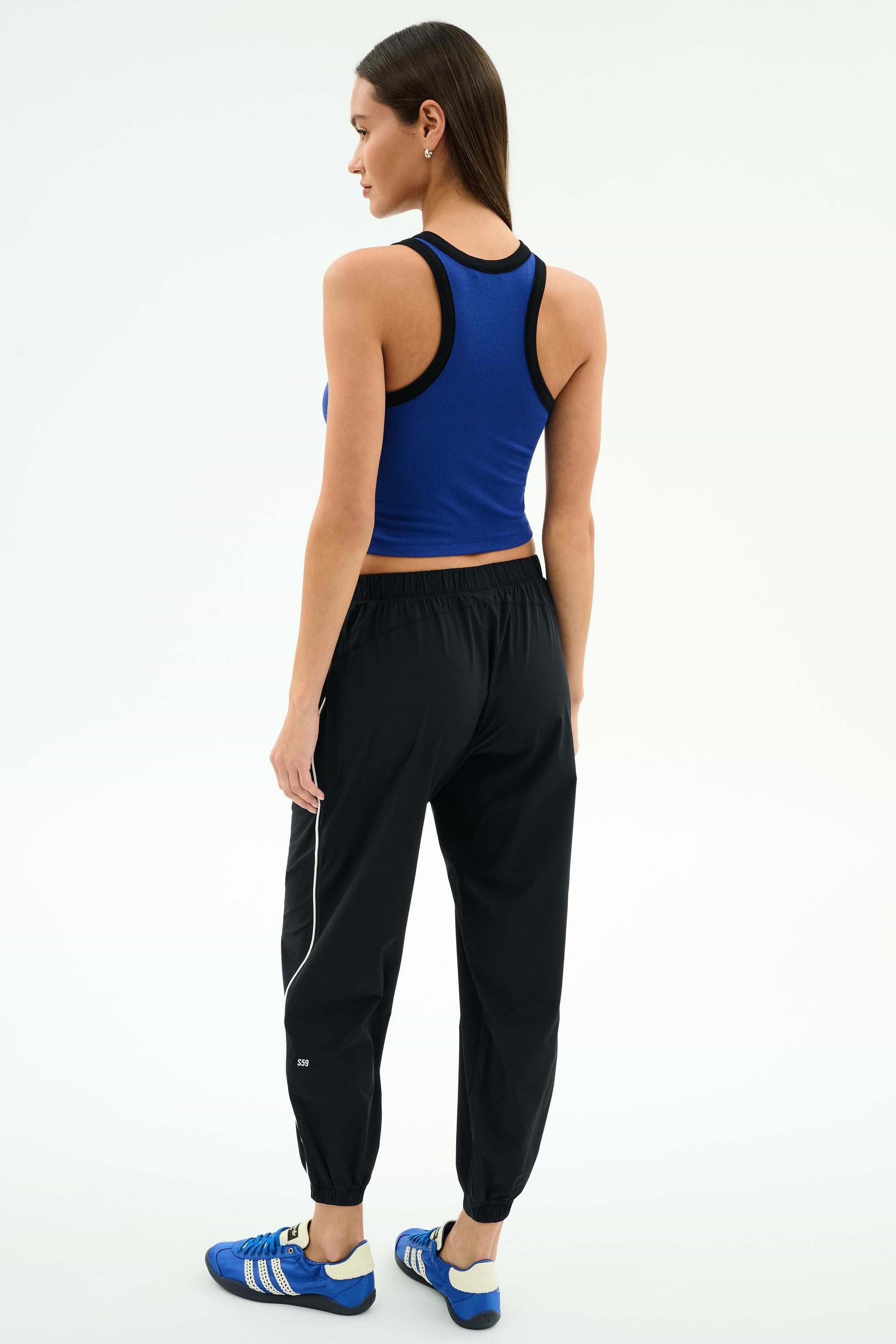 A woman faces away, wearing SPLITS59’s Megan Woven Nylon Jogger w/ Piping in Black/Creme with a blue sleeveless top and blue athletic shoes, crafted from stretchy spandex and 85% nylon, on a plain white background.