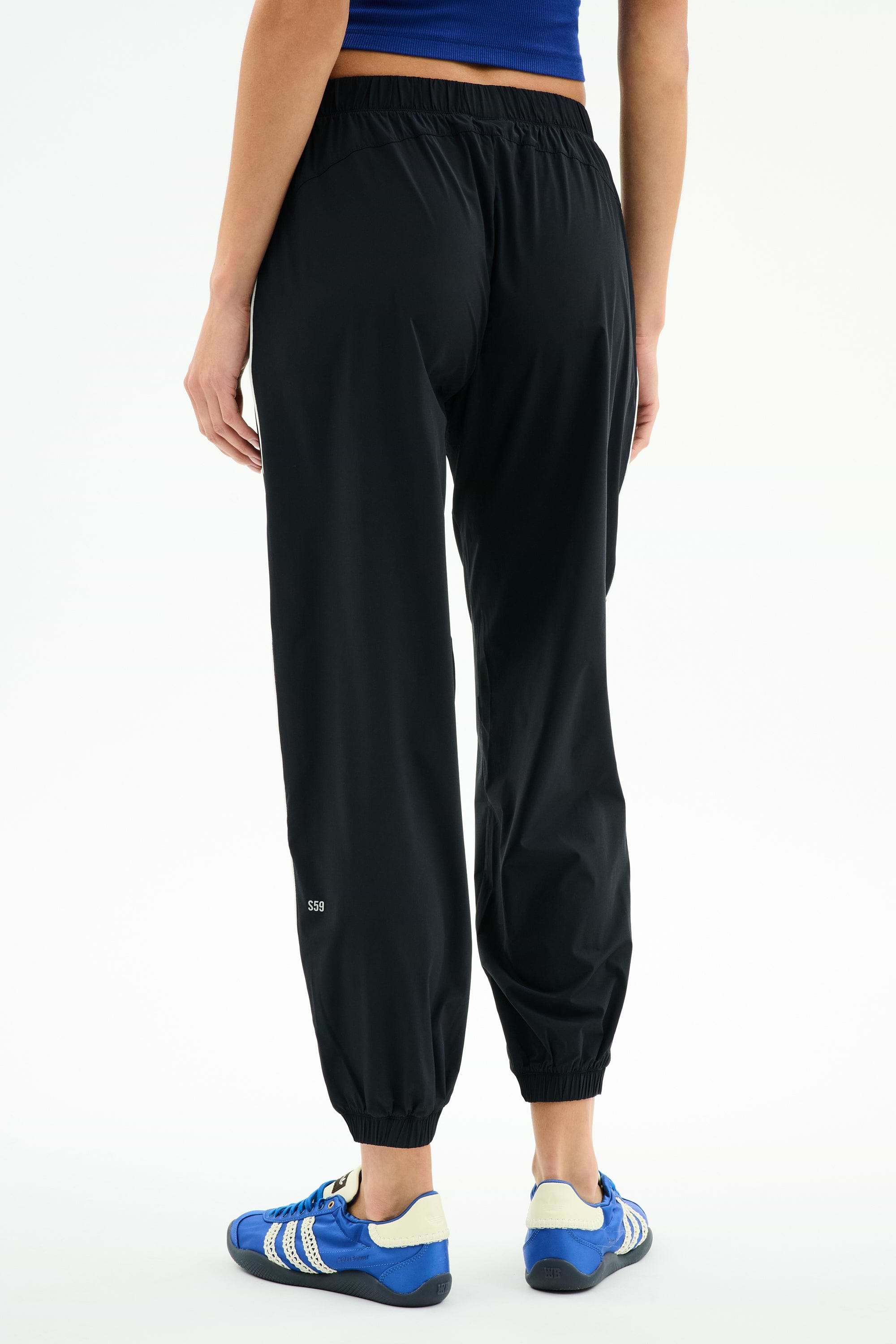 A person wears SPLITS59 Megan Woven Nylon Jogger w/ Piping - Black/Creme, a blue top, and blue athletic shoes, photographed from the back against a plain white background.