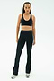 A person stands facing forward against a plain light background, wearing a black sports bra, SPLITS59 Raquel High Waist Flared Legging in Black/Creme with side stripes, and gray sneakers.