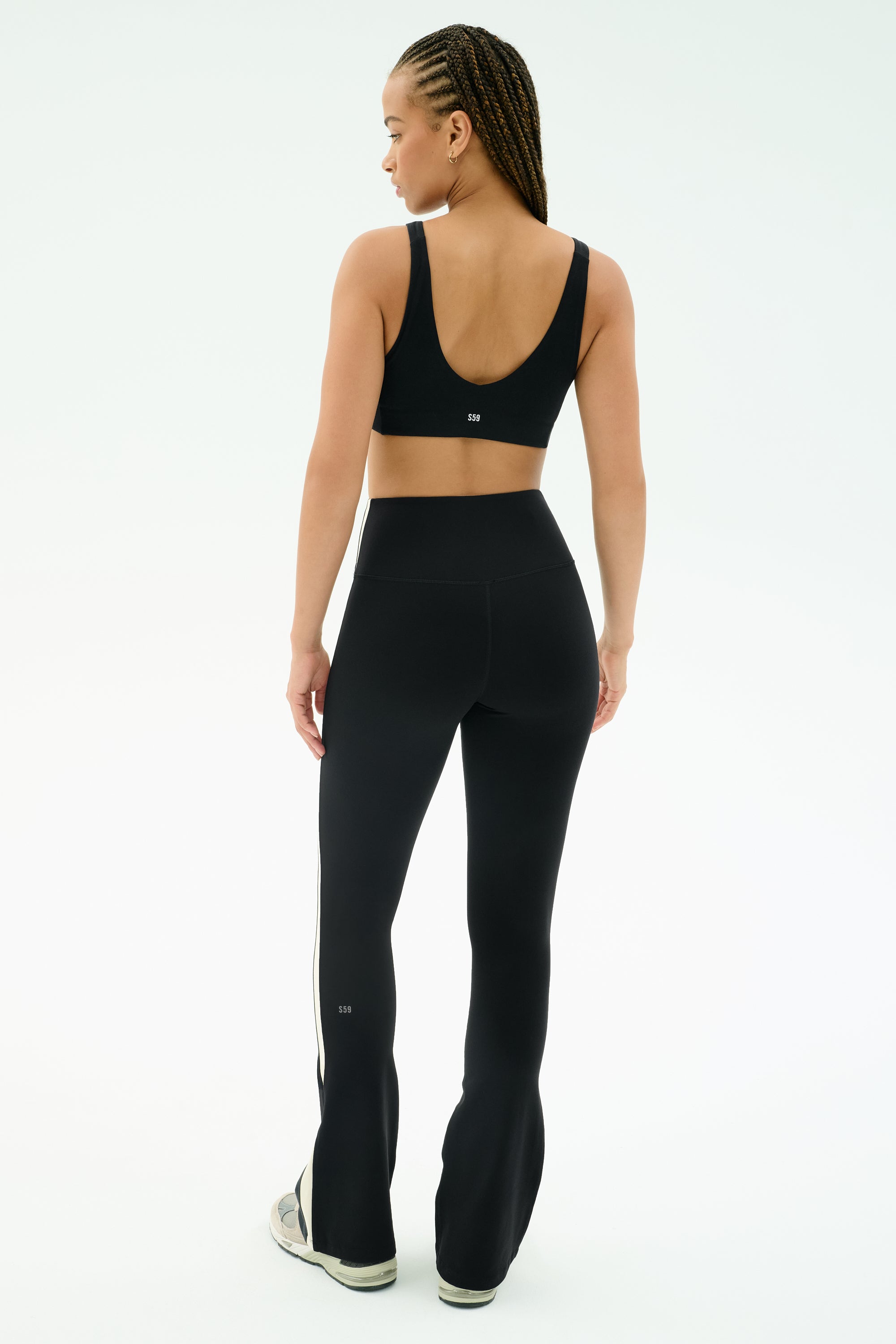 A woman faces away from the camera in SPLITS59’s Raquel High Waist Flared Legging in Black/Creme, paired with a sports bra and sneakers. She poses against a plain white background.