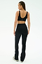 A woman faces away from the camera in SPLITS59’s Raquel High Waist Flared Legging in Black/Creme, paired with a sports bra and sneakers. She poses against a plain white background.