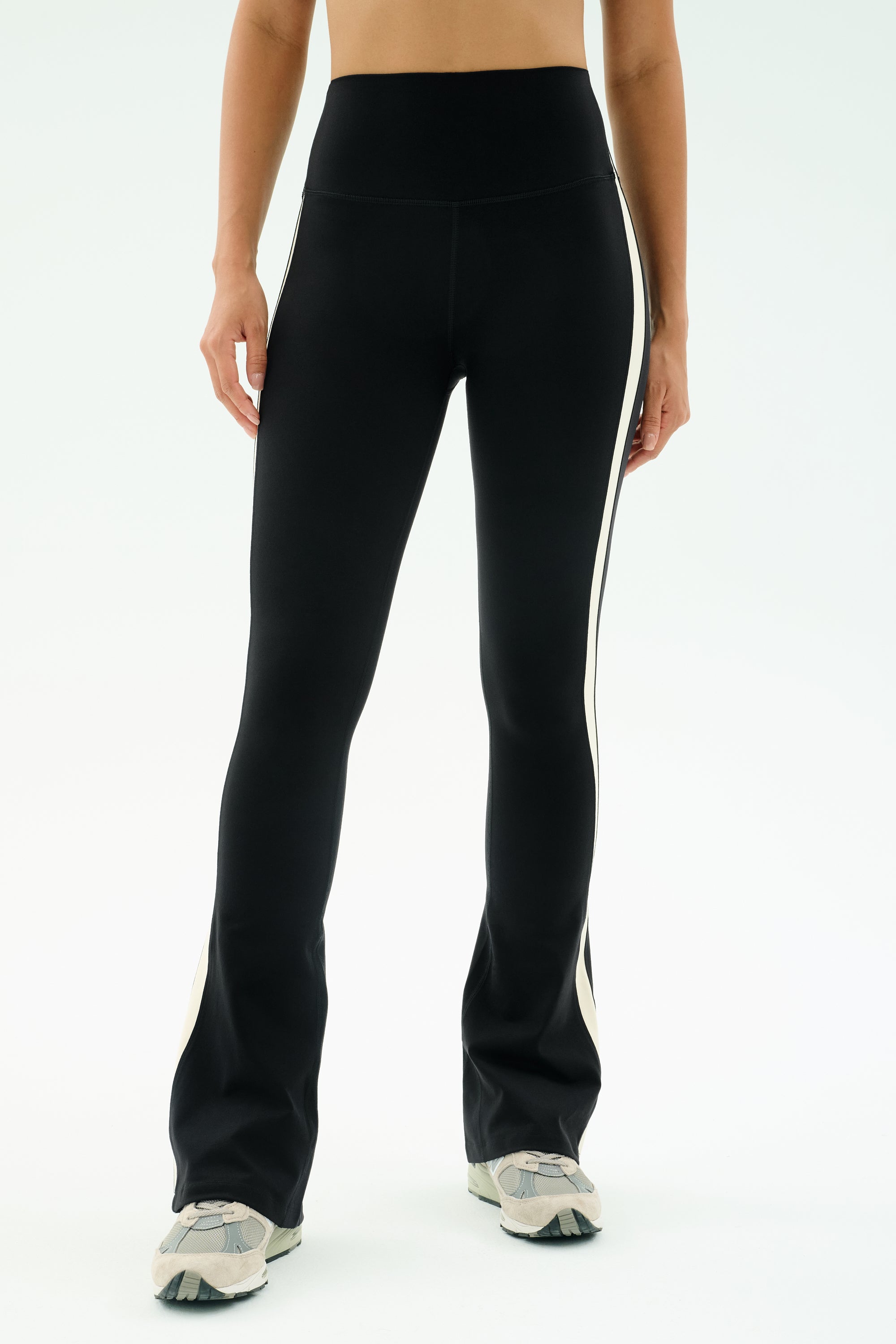 A person wears SPLITS59 Raquel High Waist Flared Legging in Black/Creme with white sneakers, standing against a plain white background.